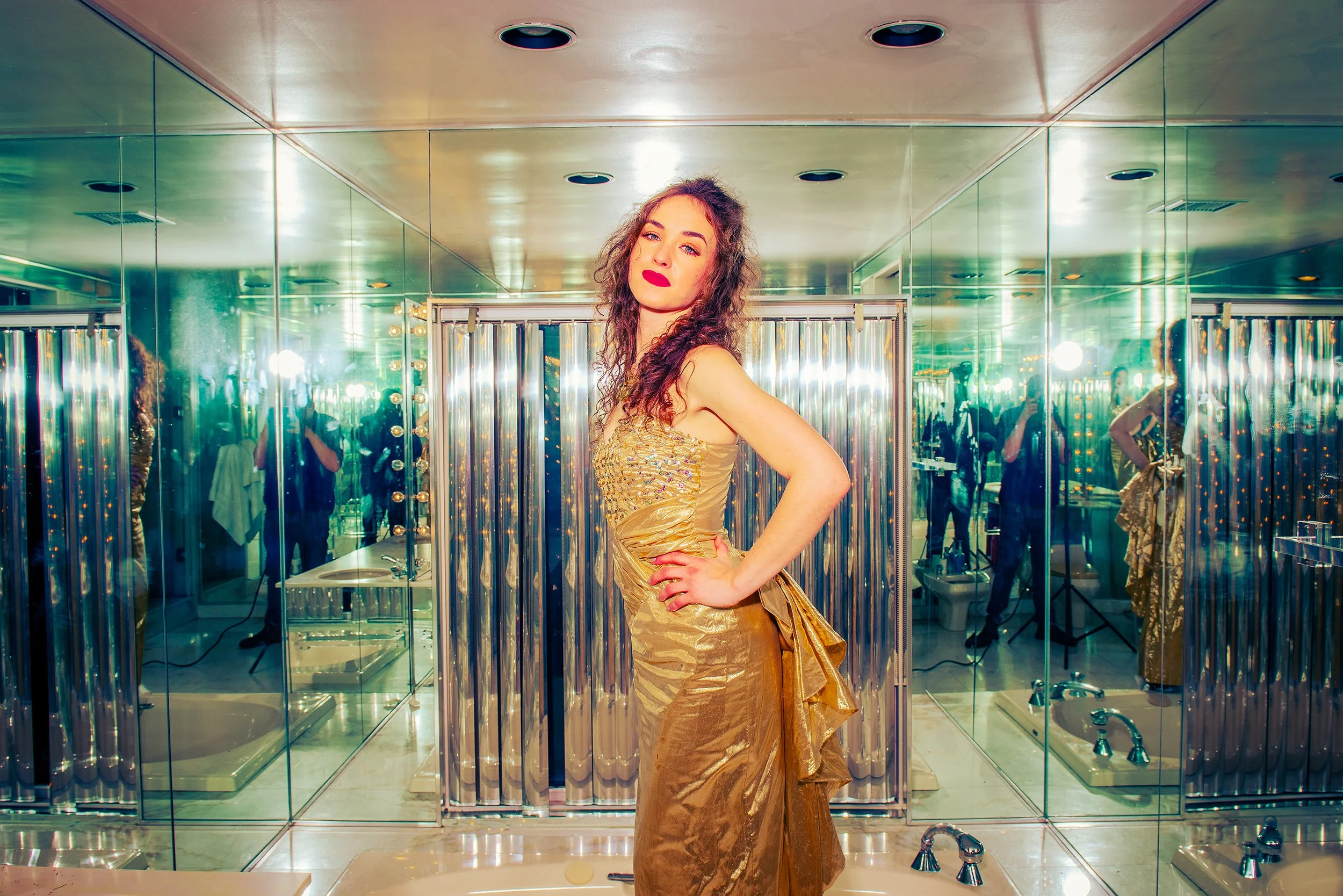A woman with wavy hair, wearing a gold dress, stands in front of mirrored walls in a bathroom, striking a pose with one hand on her hip.