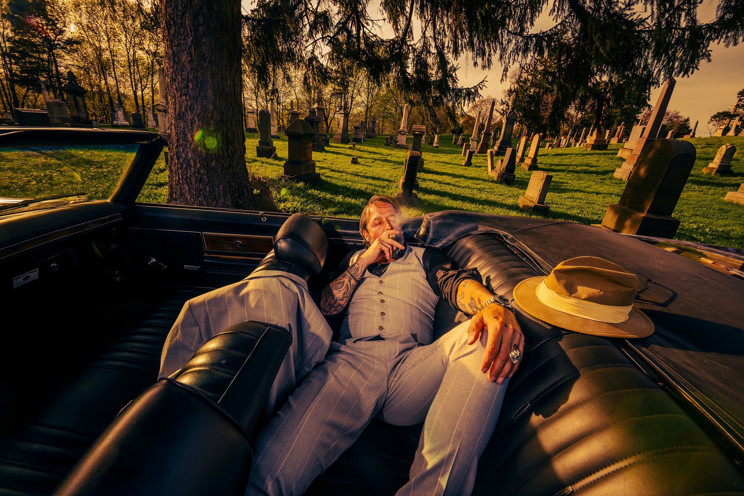 A man with tattoos relaxing in the backseat of a vintage car, smoking a cigar, with a wide-brimmed hat on his lap, set in a graveyard at sunset.