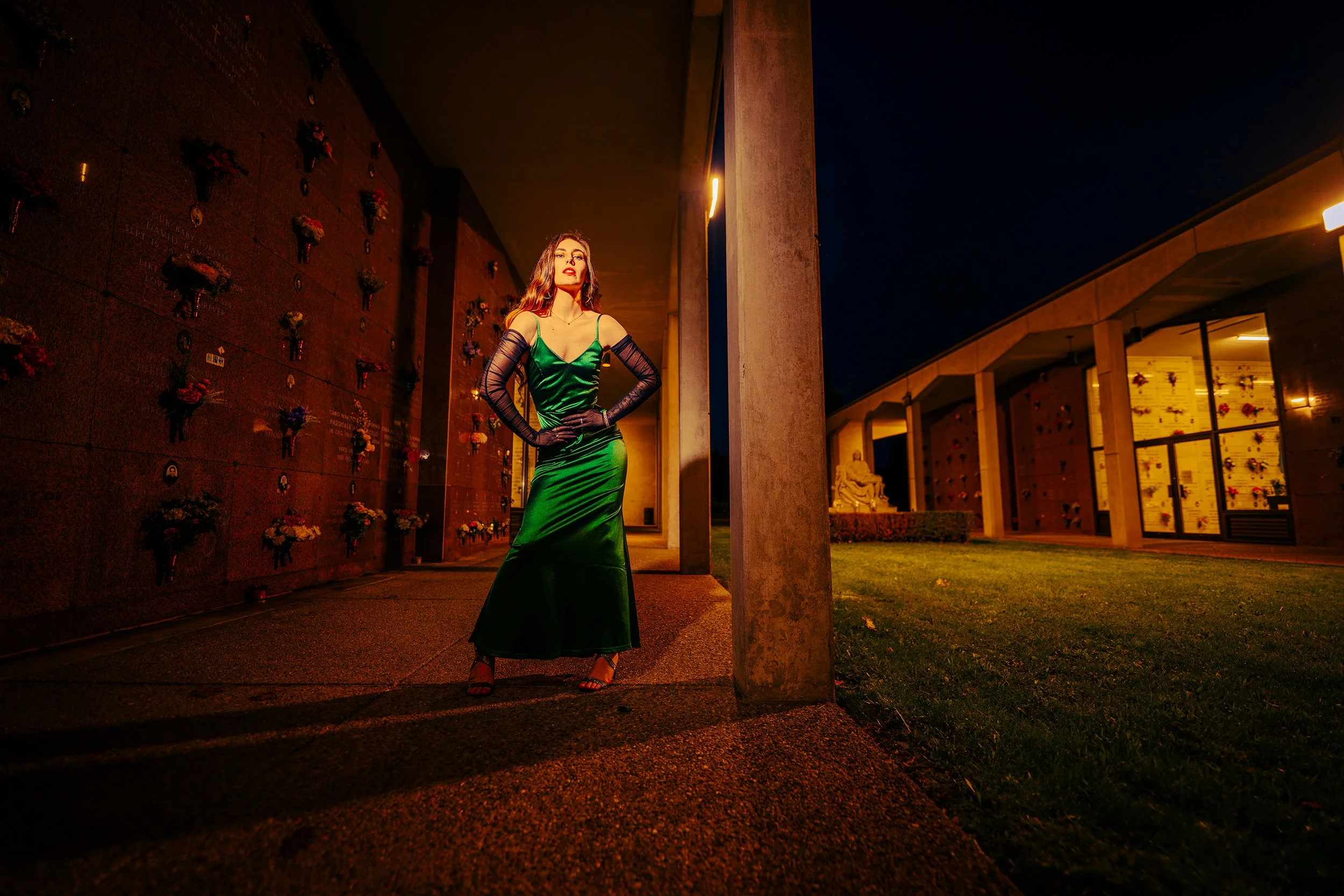 A woman in a green evening gown and black gloves stands outside at night near a memorial wall with flowers. She has long hair and red lipstick, with hands on hips, urban architecture, and a dark sky behind her.