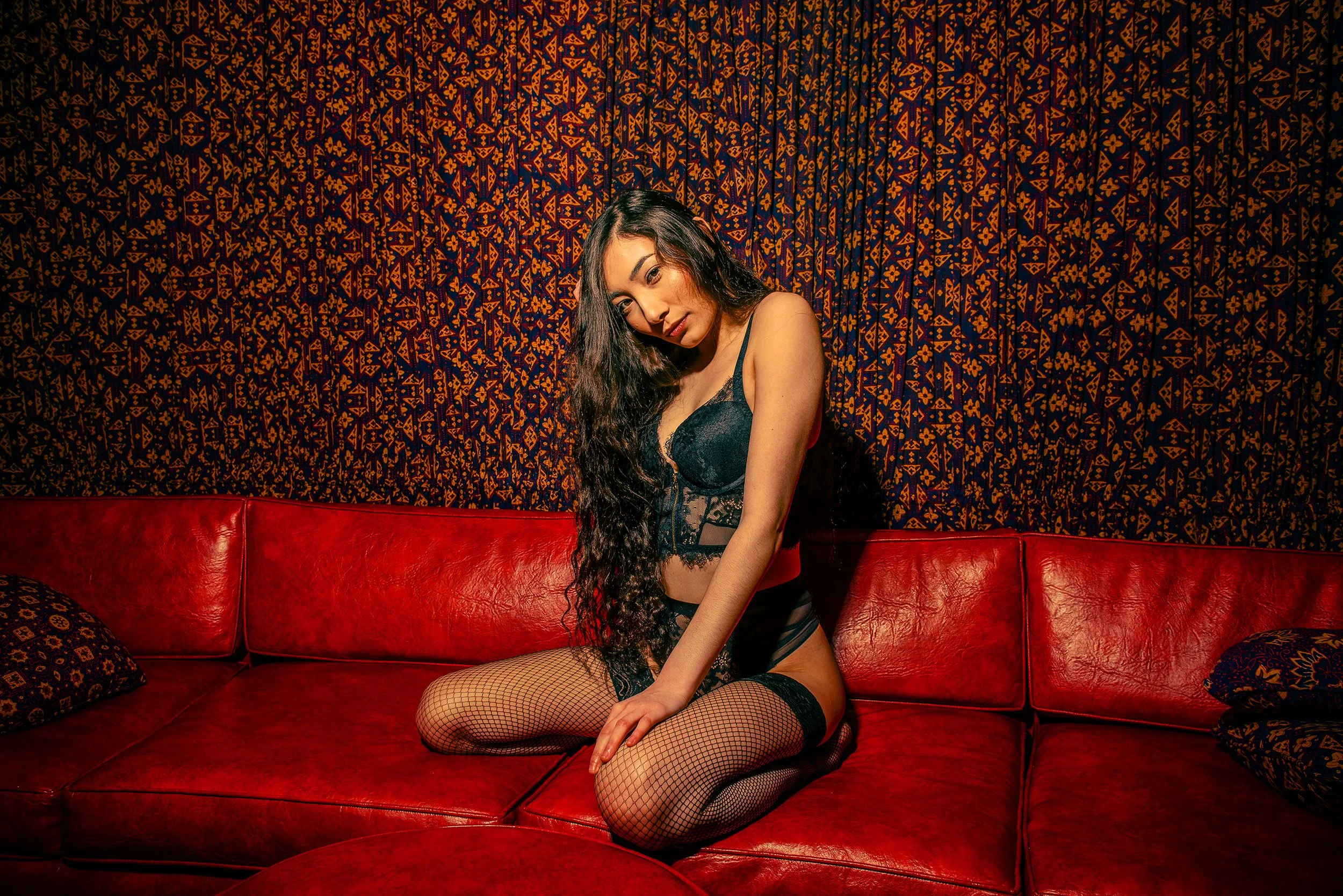 A woman with long dark hair in lingerie and fishnet stockings sitting on a red leather couch in front of a patterned wall.
