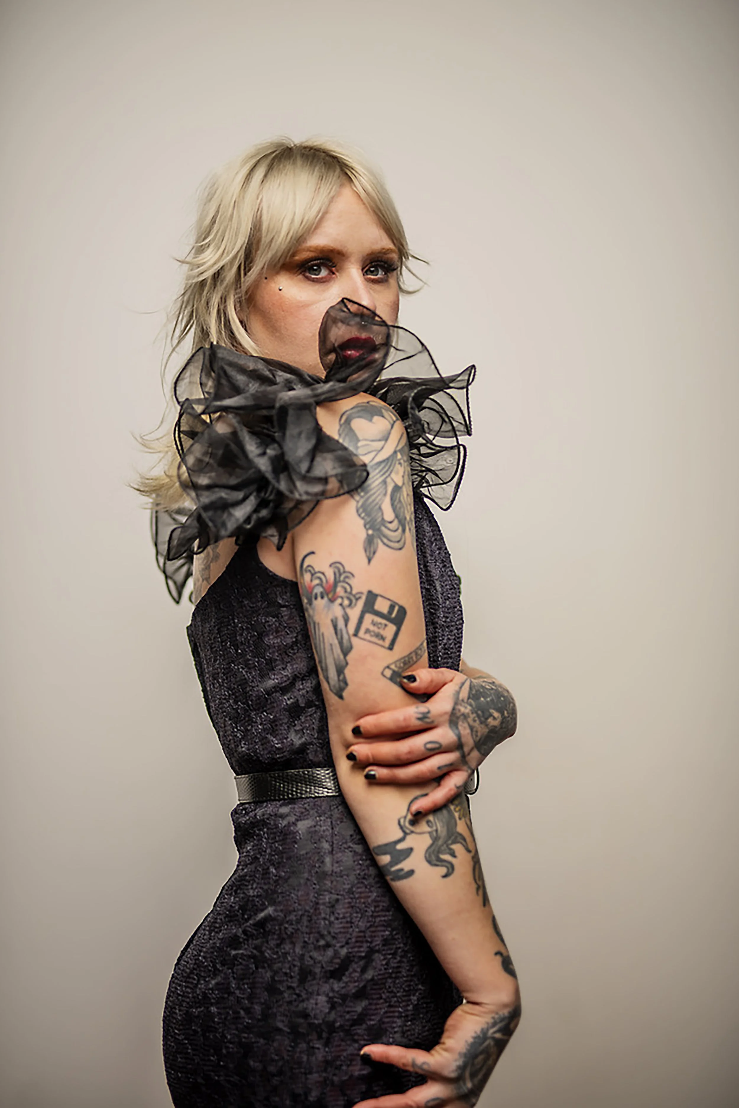 A woman with blonde hair and tattoos, wearing a dark dress with sheer ruffled sleeves, is posing against a plain background with her arms crossed.