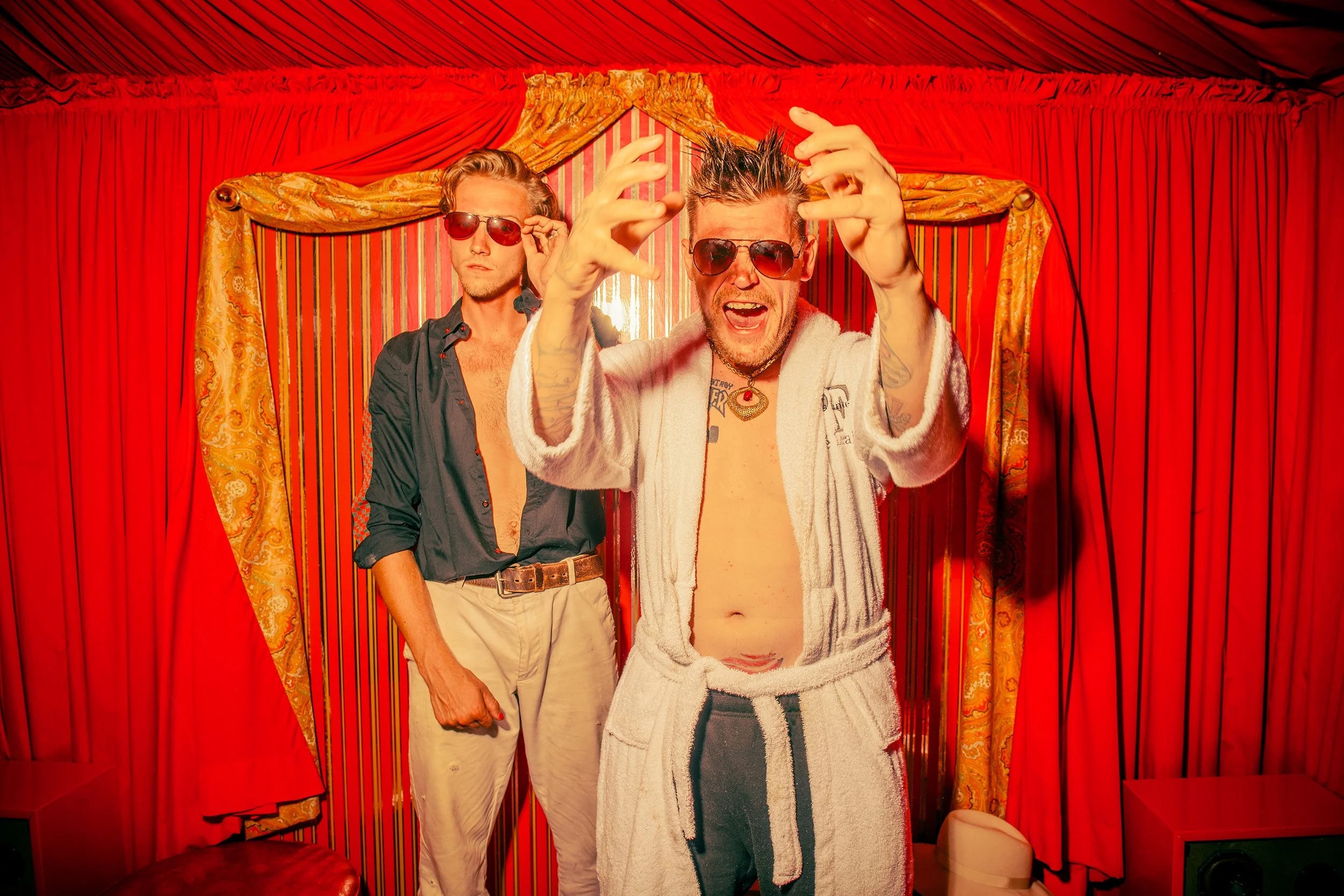 Two men with sunglasses posing in front of red curtains and gold drapes, one wearing a towel and the other a dark shirt with rolled-up sleeves.