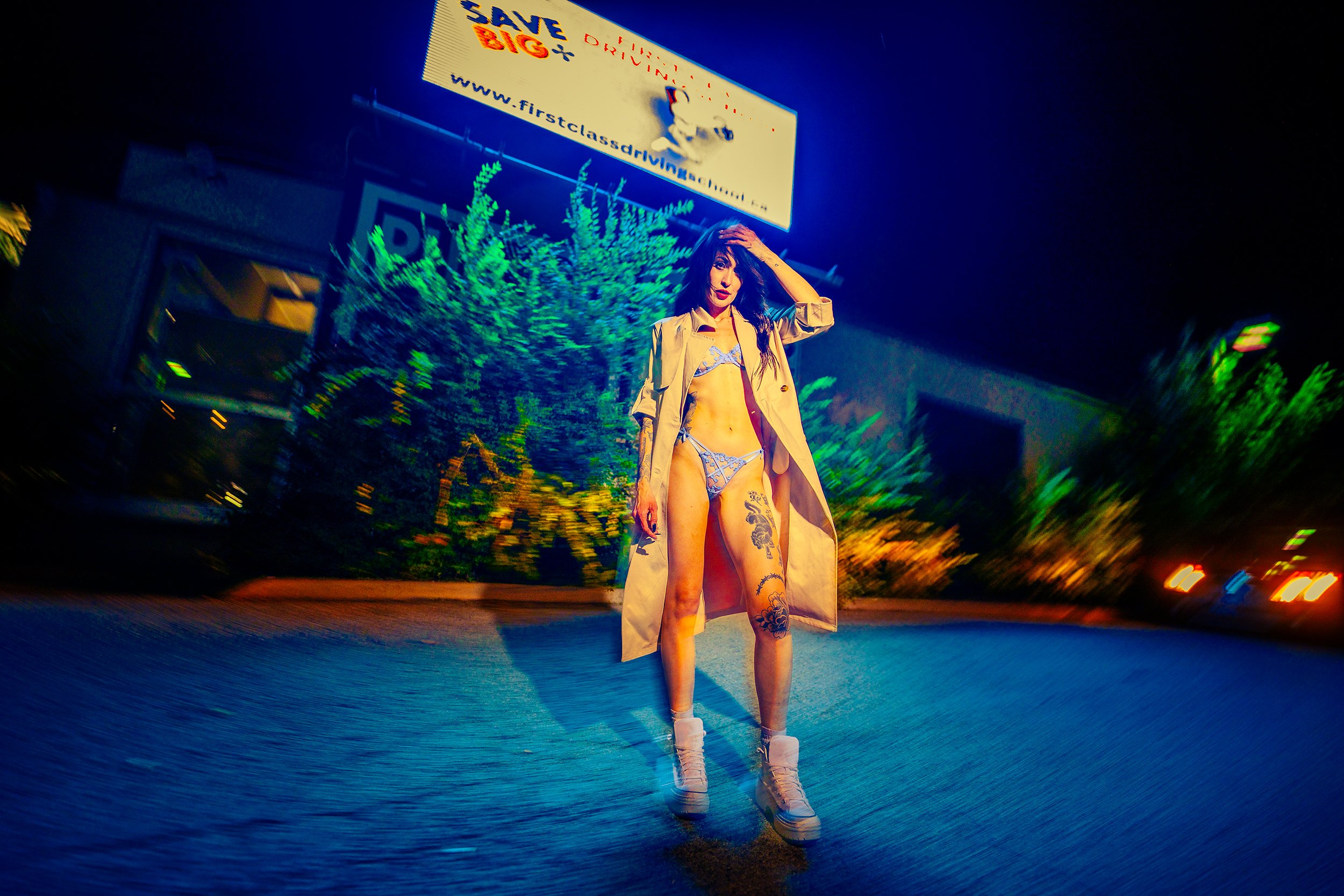 A woman stands on a street at night wearing lingerie and a trench coat, with one hand on her head, illuminated by colorful neon lights and a billboard above her.