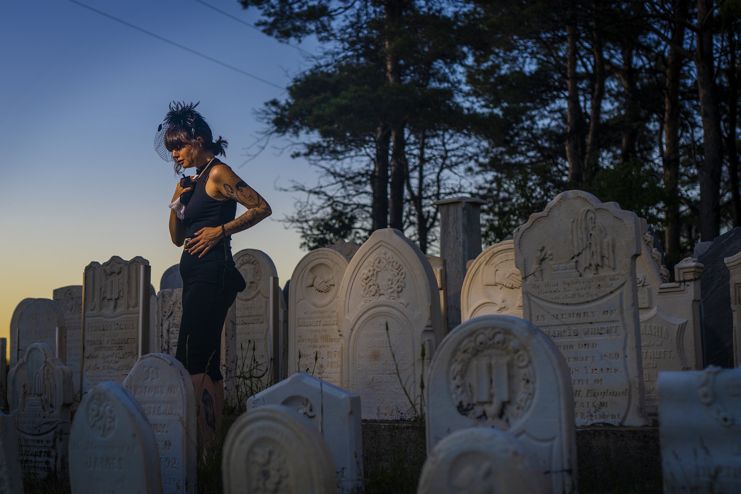 A woman dressed in black with tattoos on her arm, wearing a black fascinator hat with a veil, standing among gravestones in a cemetery at sunset.