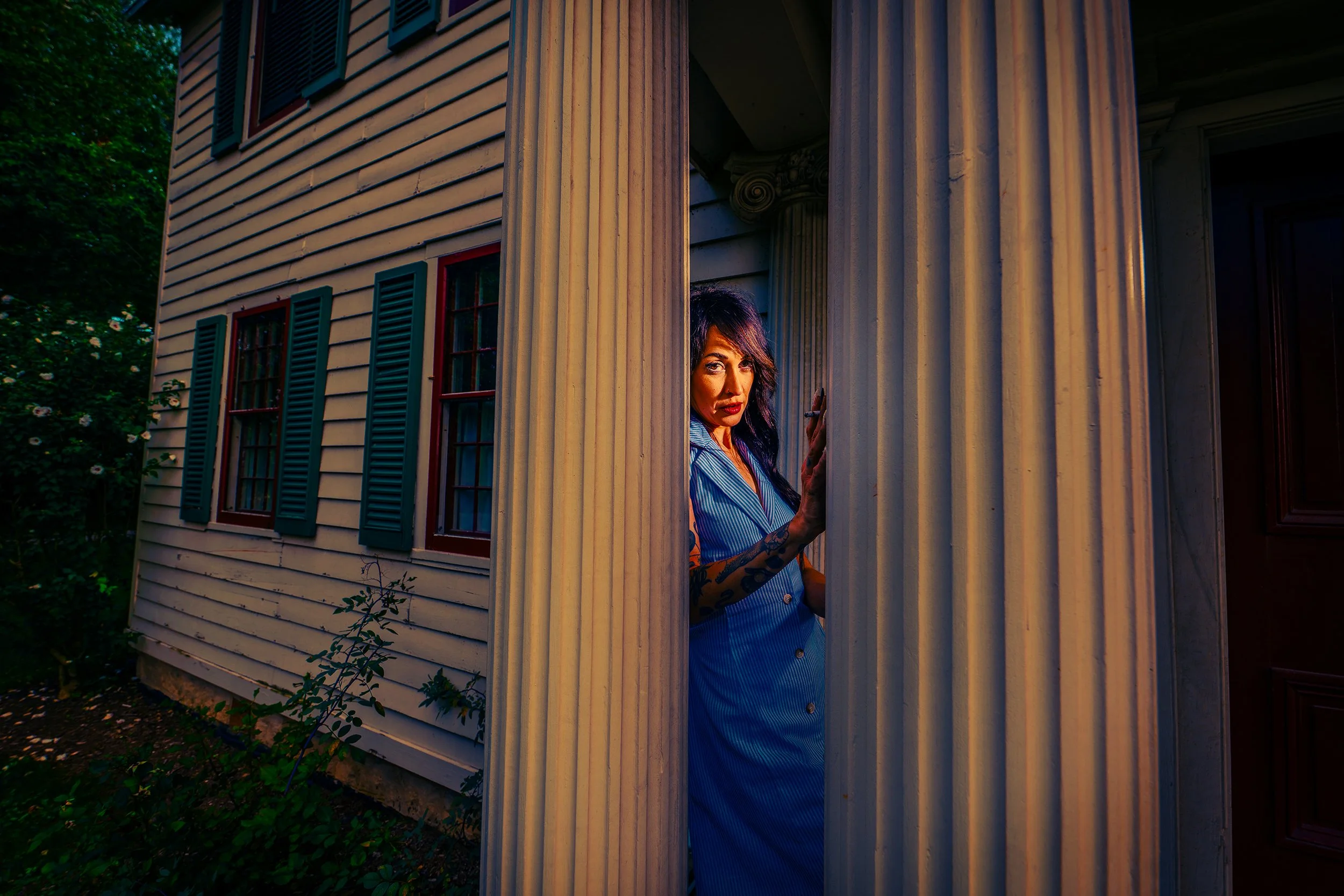 A woman standing behind a porch pillar outside a house with cream siding and dark shutters, partially visible as she peeks through the gap.
