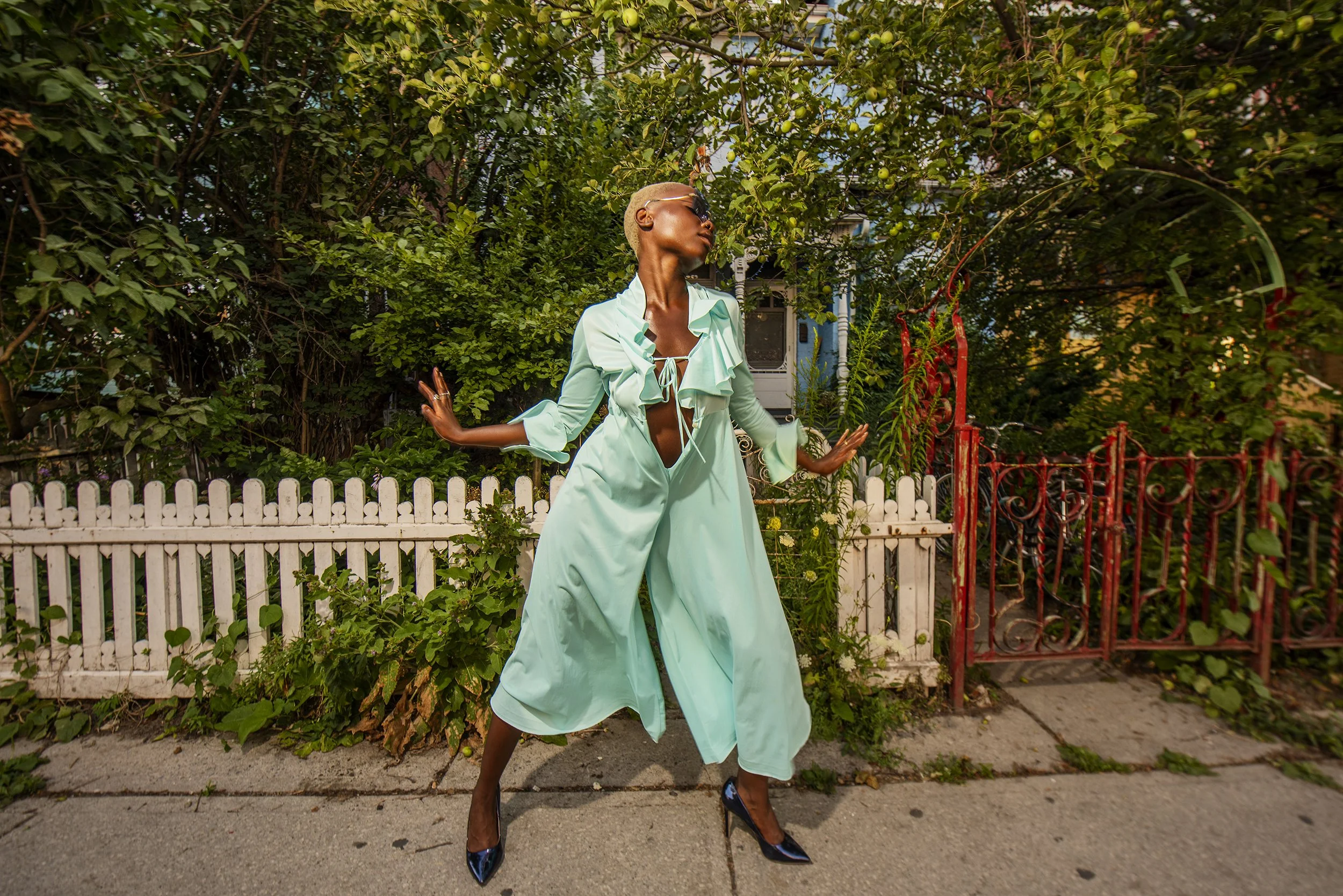 A person with short blond hair and dark skin, wearing dark sunglasses, a light green blouse with ruffles and tie in front, matching long skirt, and black high heels, is standing with arms outstretched in front of a lush green garden and white picket 