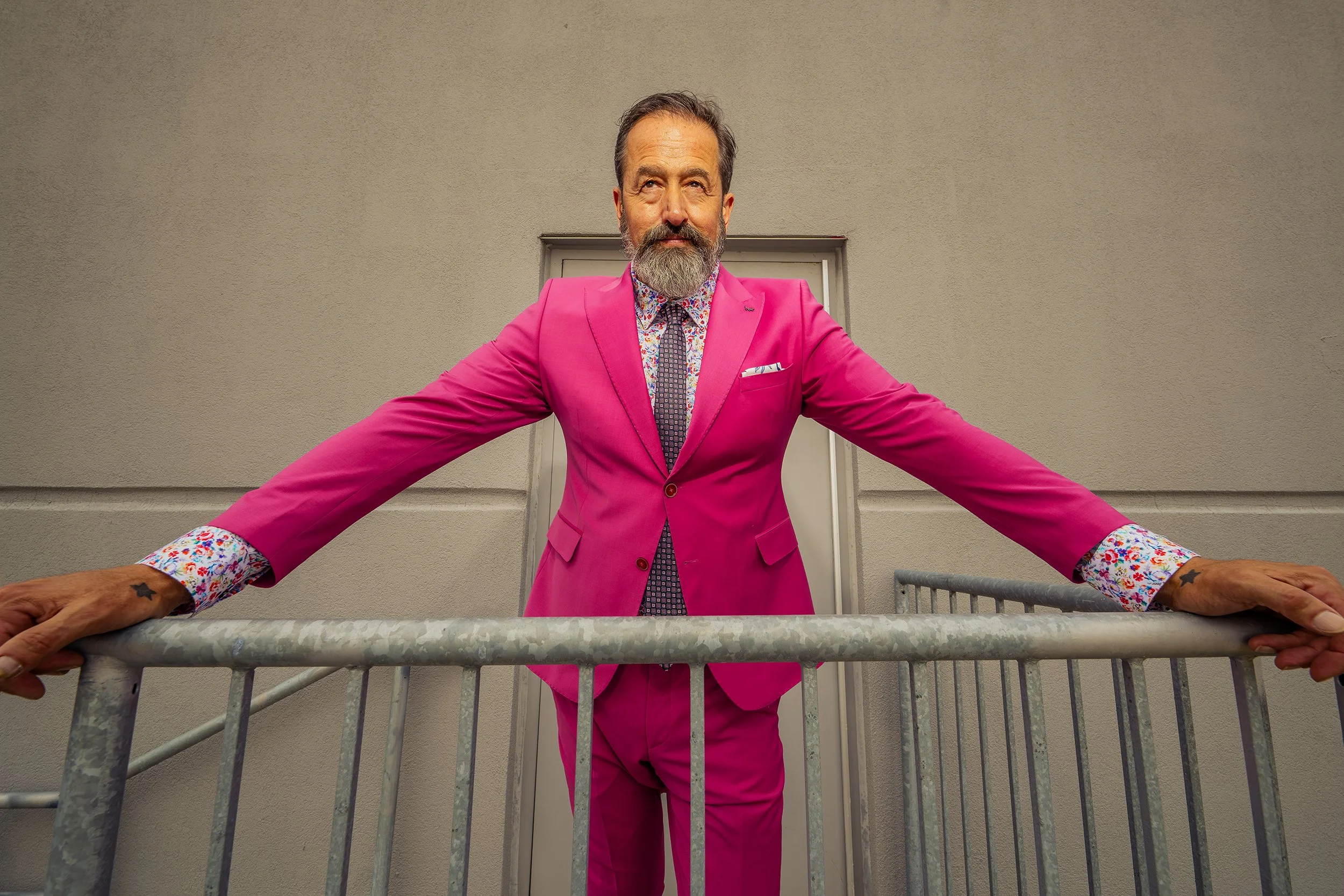 A man stands on a balcony wearing a bright pink suit with a floral shirt and a patterned tie, holding onto the railing with outstretched arms.