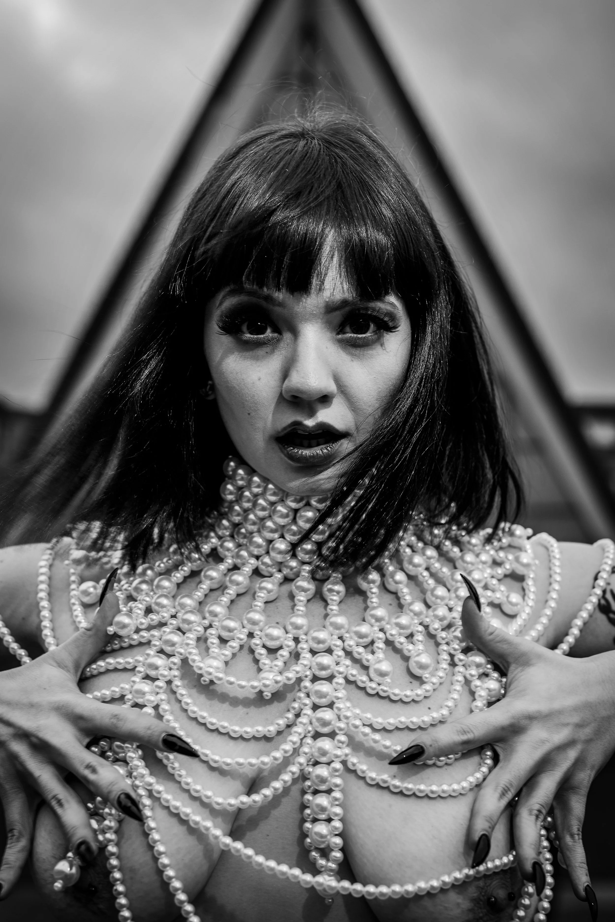 A woman with dark hair and bangs, holding layers of pearl necklaces and beads against her chest, in front of a mirror with an A-frame reflection, in black and white.