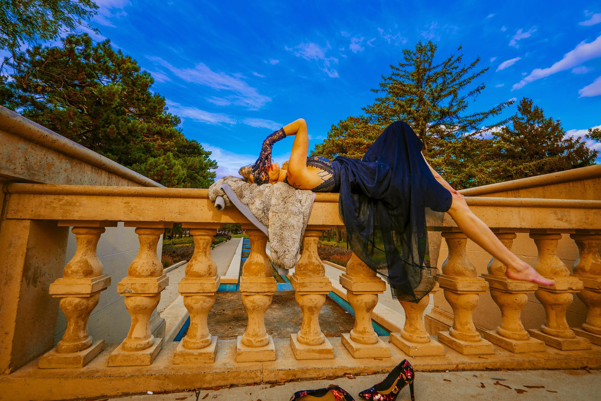 Woman lying on a stone railing with her eyes closed, dressed in a black lace dress, with colorful high heels and a jacket nearby, outdoors under a blue sky with trees in the background.
