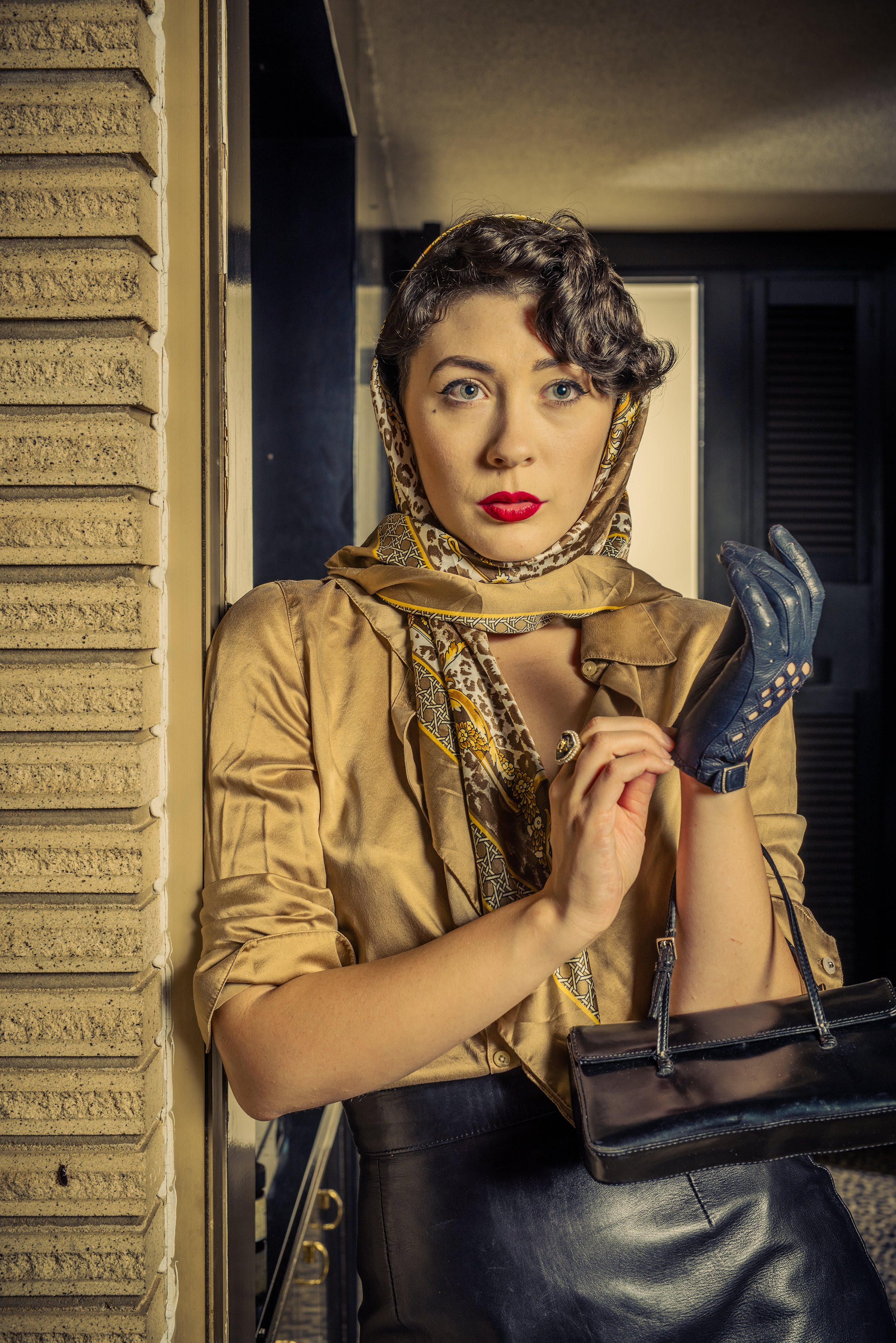 A woman with short dark curly hair and blue eyes, wearing bright red lipstick, dressed in a shiny brown jacket, patterned silk scarf, and black leather skirt, holding a black handbag, standing on a city sidewalk leaning against a brick wall, with a n