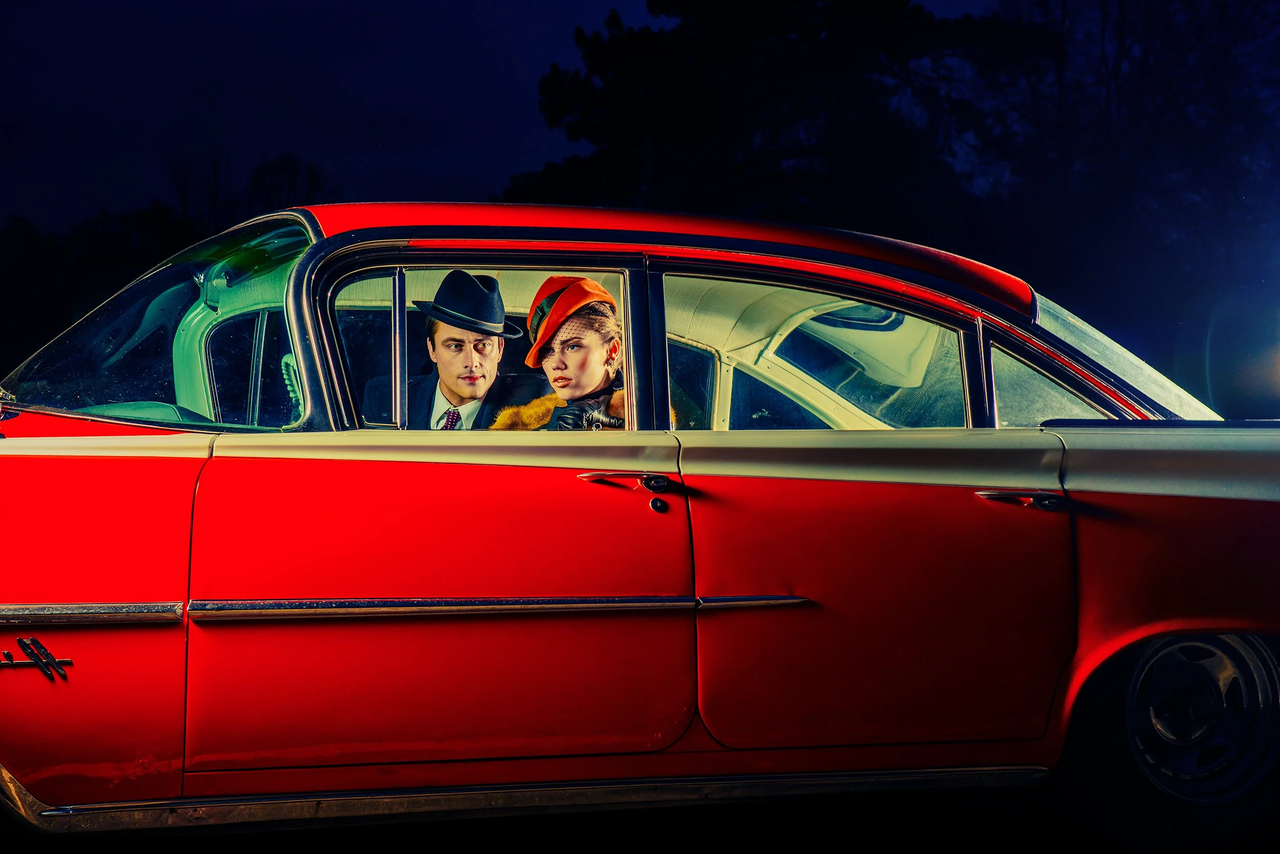 A man and woman sitting inside a vintage red and black car at night. The man is wearing a black fedora and a suit, while the woman is wearing an orange hat with green details and a fur coat. They are looking out the car window.