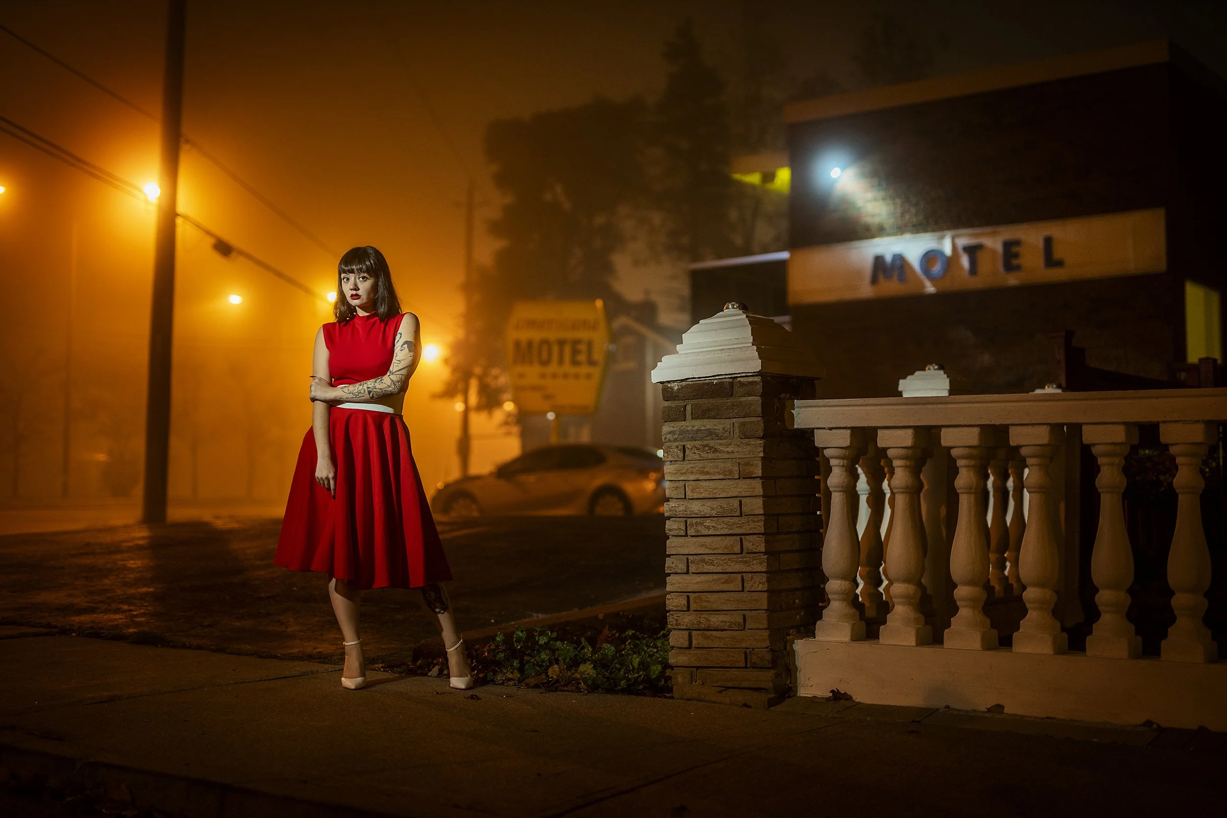 A woman in a red dress with tattoos on her arm and leg, standing on a foggy street at night with orange streetlights, near a motel sign and building.