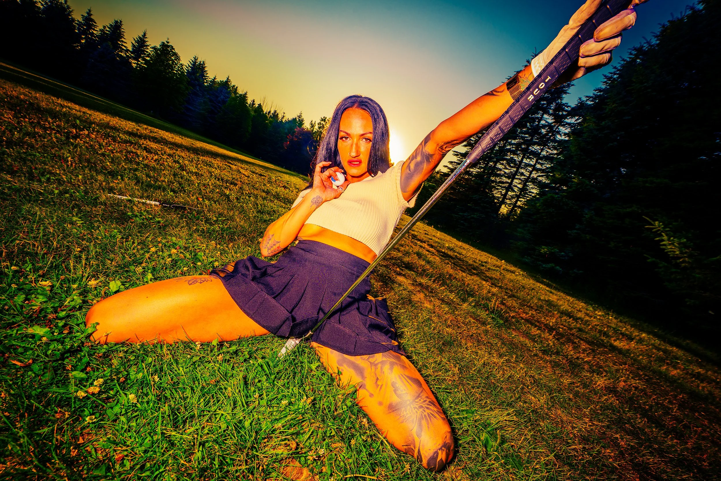 A woman with tattoos, dark hair, and wearing a cream crop top and black shorts, holding a golf club while sitting on a grassy field during sunset.