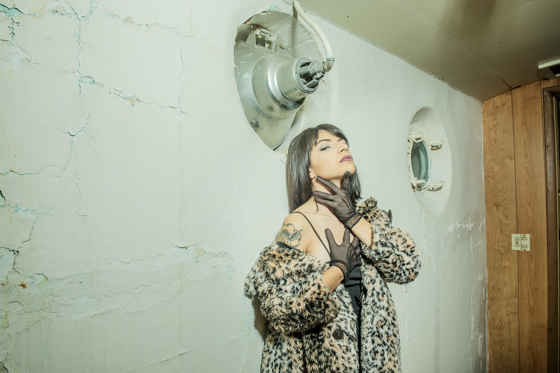 A woman wearing a leopard print coat, black gloves, and black top, poses against a cracked white wall with round porthole windows and an industrial pipe above her head.