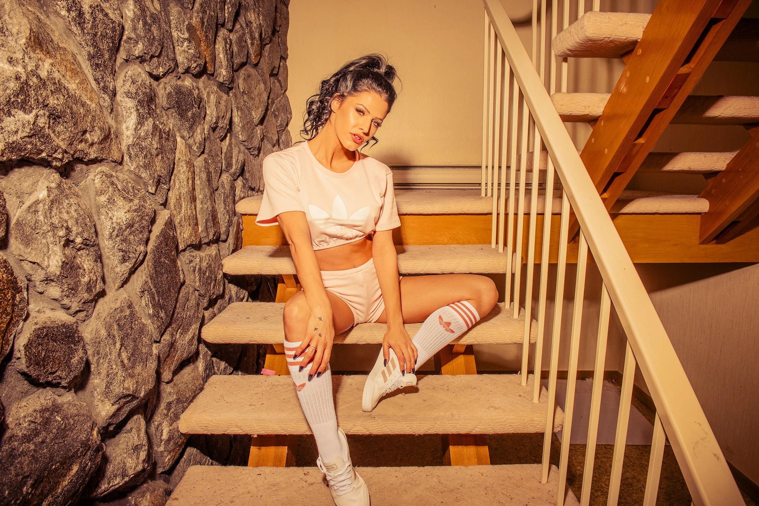 A woman sitting on carpeted stairs, leaning against a stone wall, wearing a pink crop top with the Adidas logo, pink shorts, knee-high white socks with red stripes, and white sneakers, with curly dark hair and makeup.