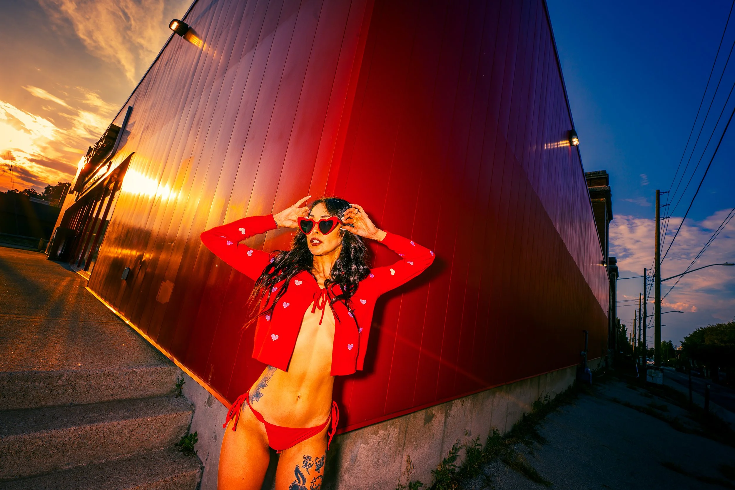 A woman wearing heart-shaped sunglasses and a red jacket with white hearts, standing against a large red wall during sunset.