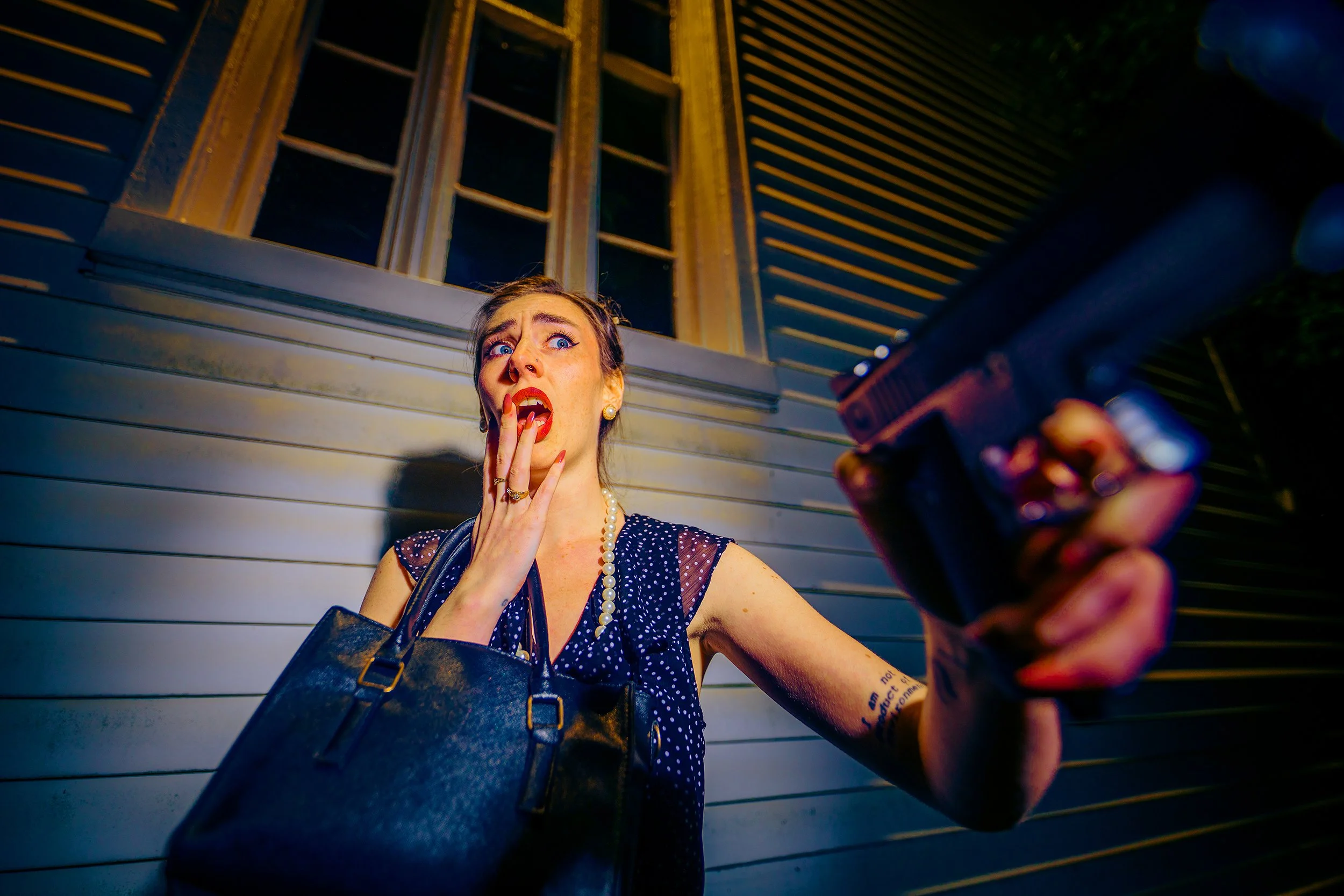 A woman with blue eyes and red lipstick, dressed in vintage clothing, looks shocked with her right hand on her mouth and left hand holding a gun pointed at her. She is standing in front of a wooden building.