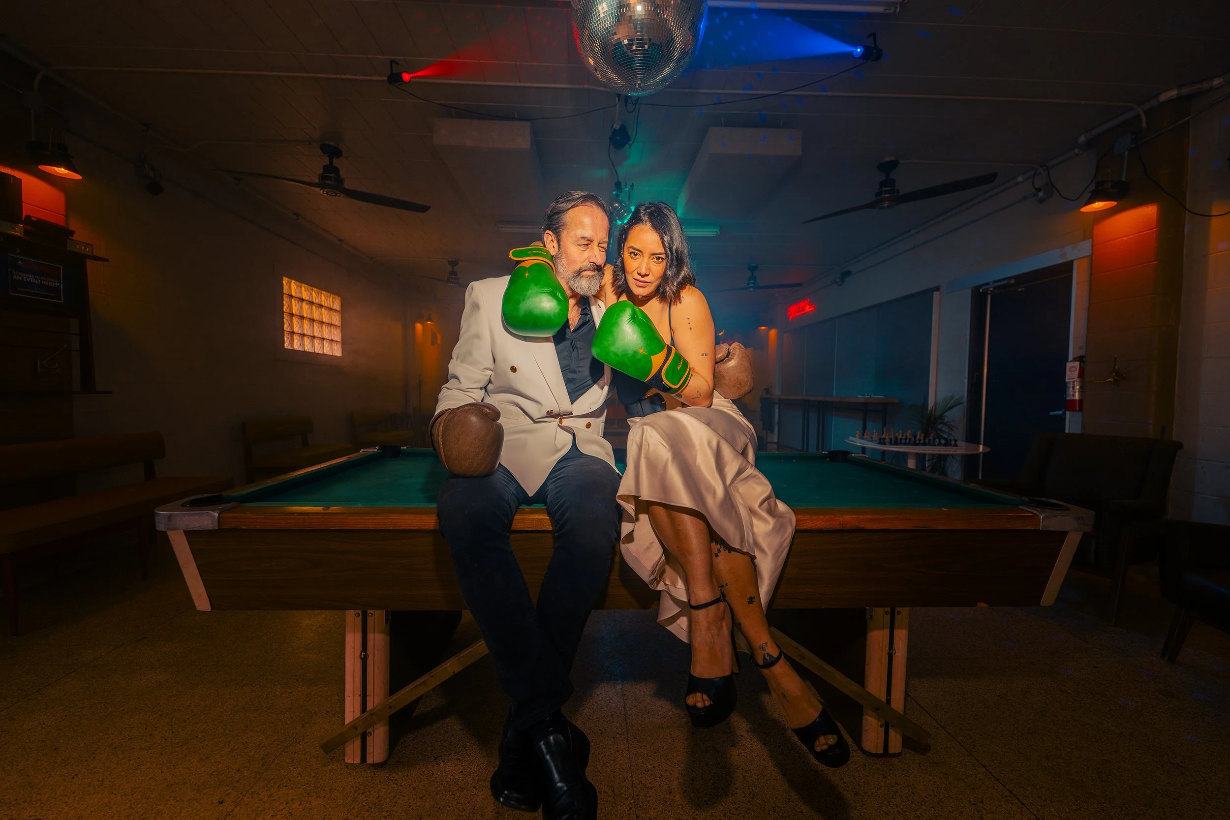 Two people sitting on a pool table, wearing boxing gloves, in a dimly lit room with colorful lights and a disco ball overhead, suggesting a party or playful setting.