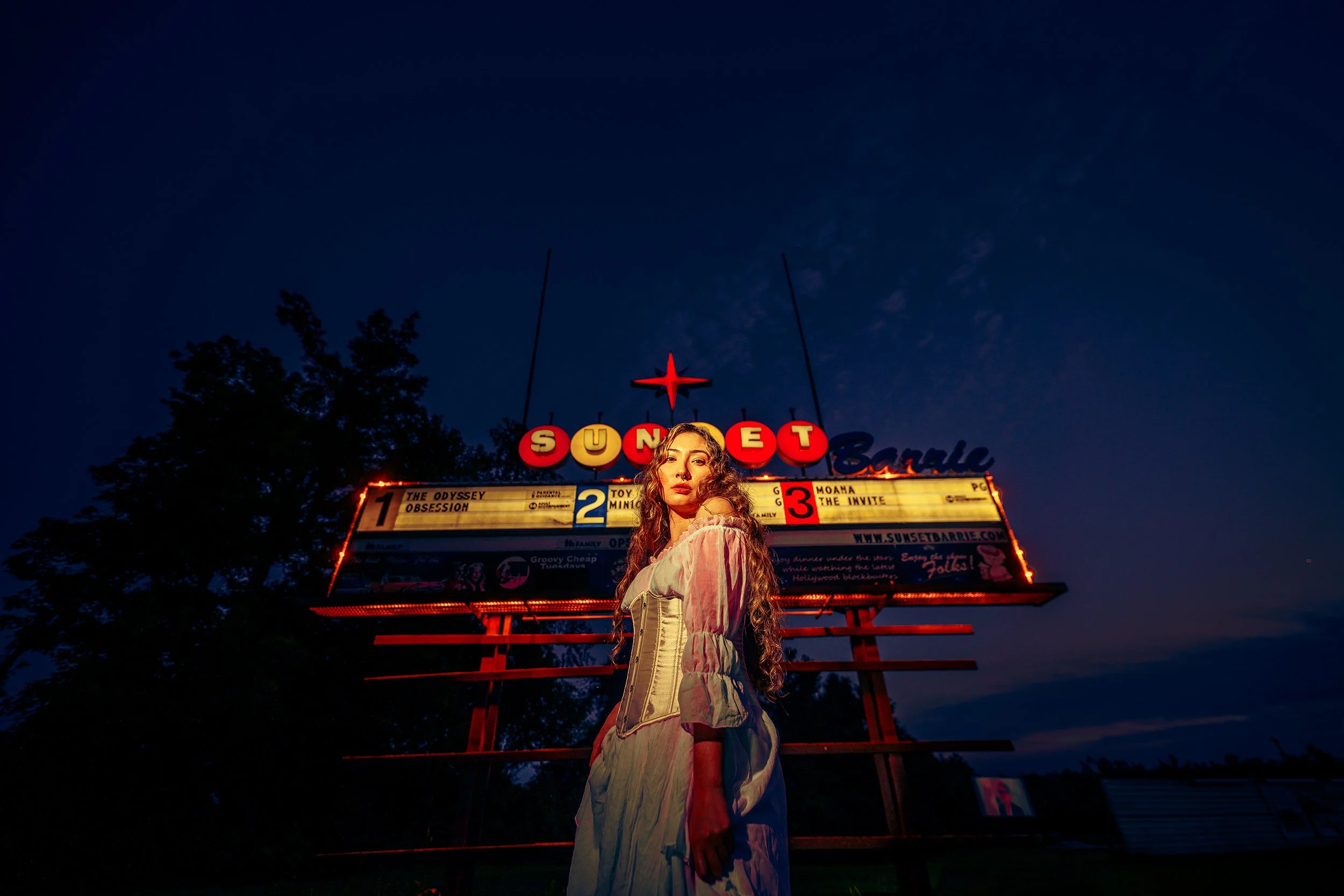 A young woman with long, curly hair standing in front of a vintage neon-lit drive-in theater marquee at dusk or night. The marquee displays movie titles and a website address, with bright red and yellow lights and a star symbol at the top. The sky is