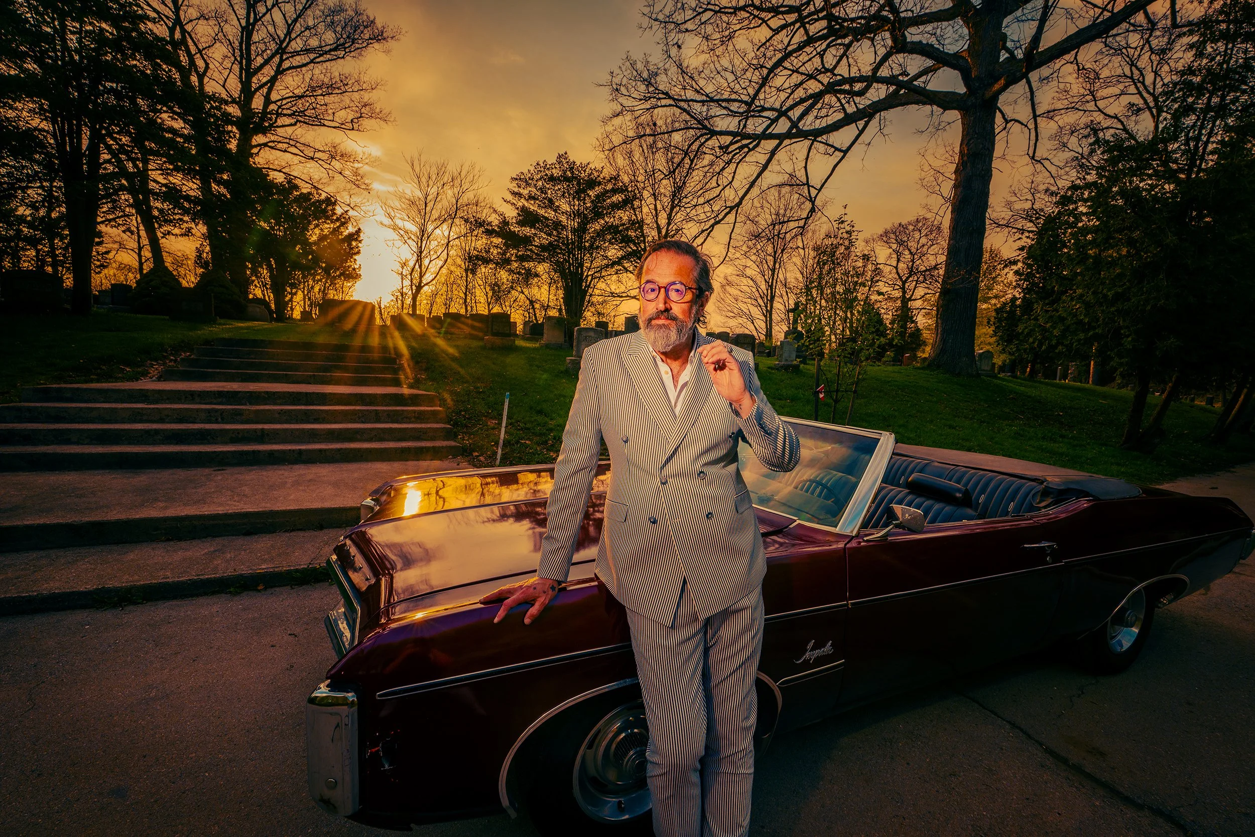 A man with glasses and a beard dressed in a striped suit standing next to a vintage convertible car during sunset in a park with trees and stairs.