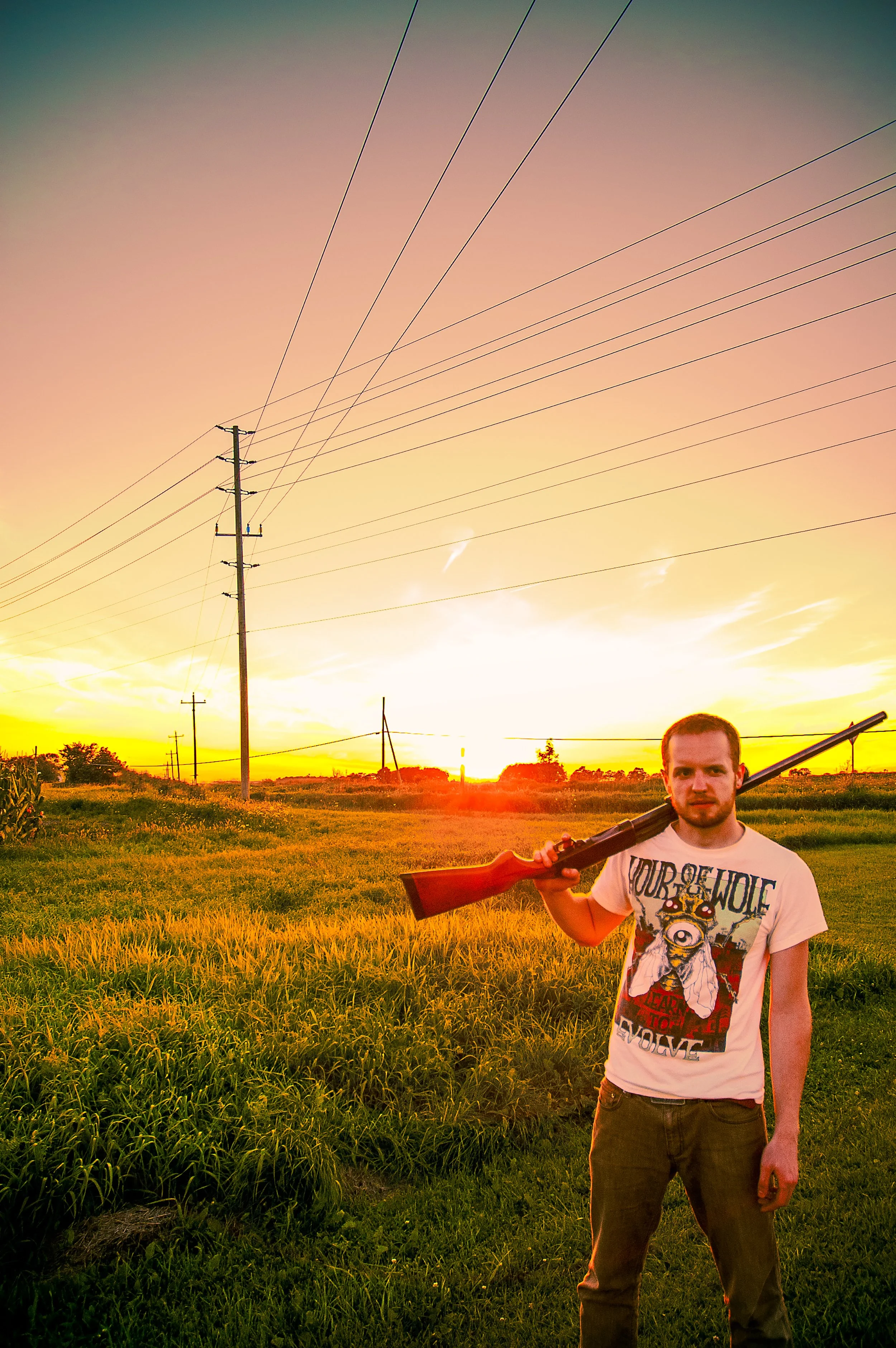 A man holding a rifle standing in a field during sunset with power lines in the background.