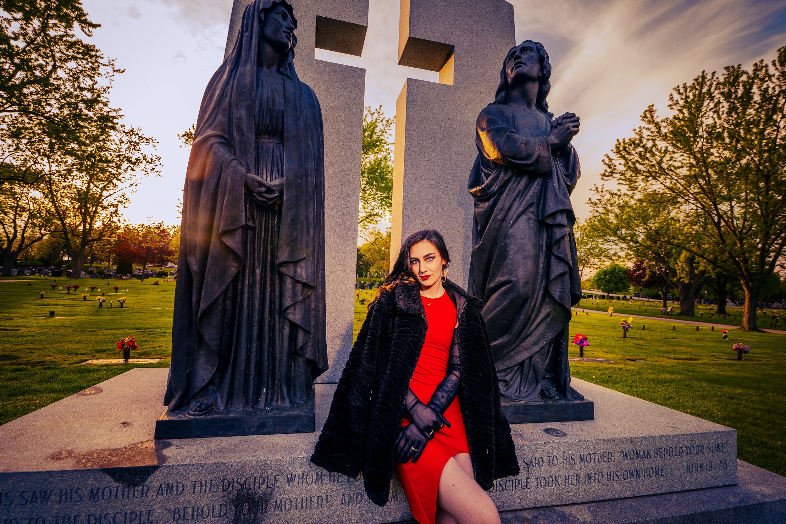 Young woman with dark hair and red dress standing between two large religious statues in a cemetery during sunset. The statues depict Mary and Jesus, with trees and graves visible in the background.