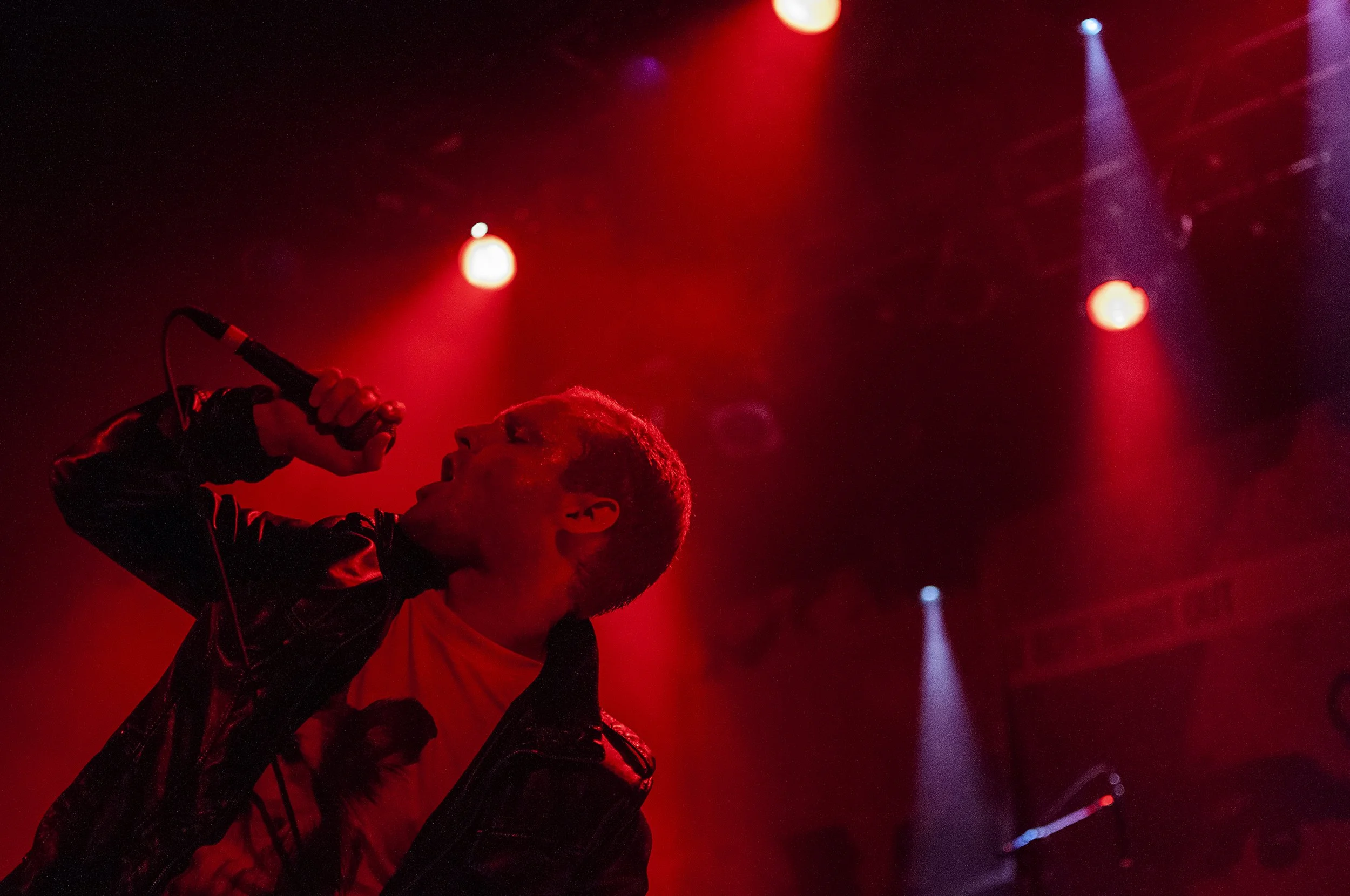 A singer performing live on stage, holding a microphone close to his mouth, with red stage lighting and visible spotlights in the background.