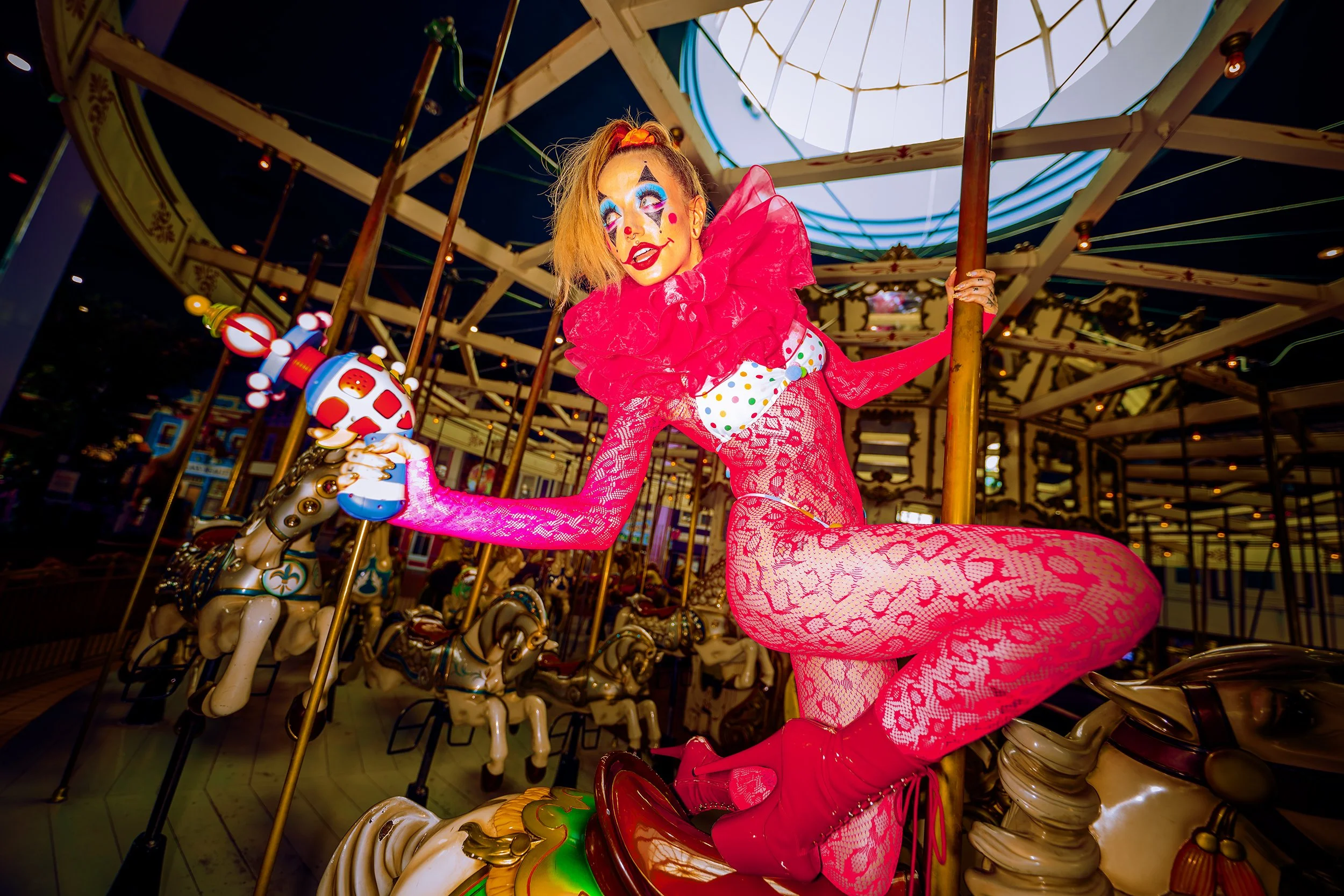 A woman dressed as a clown with colorful makeup and a bright pink dress riding a carousel horse at an amusement park during nighttime.