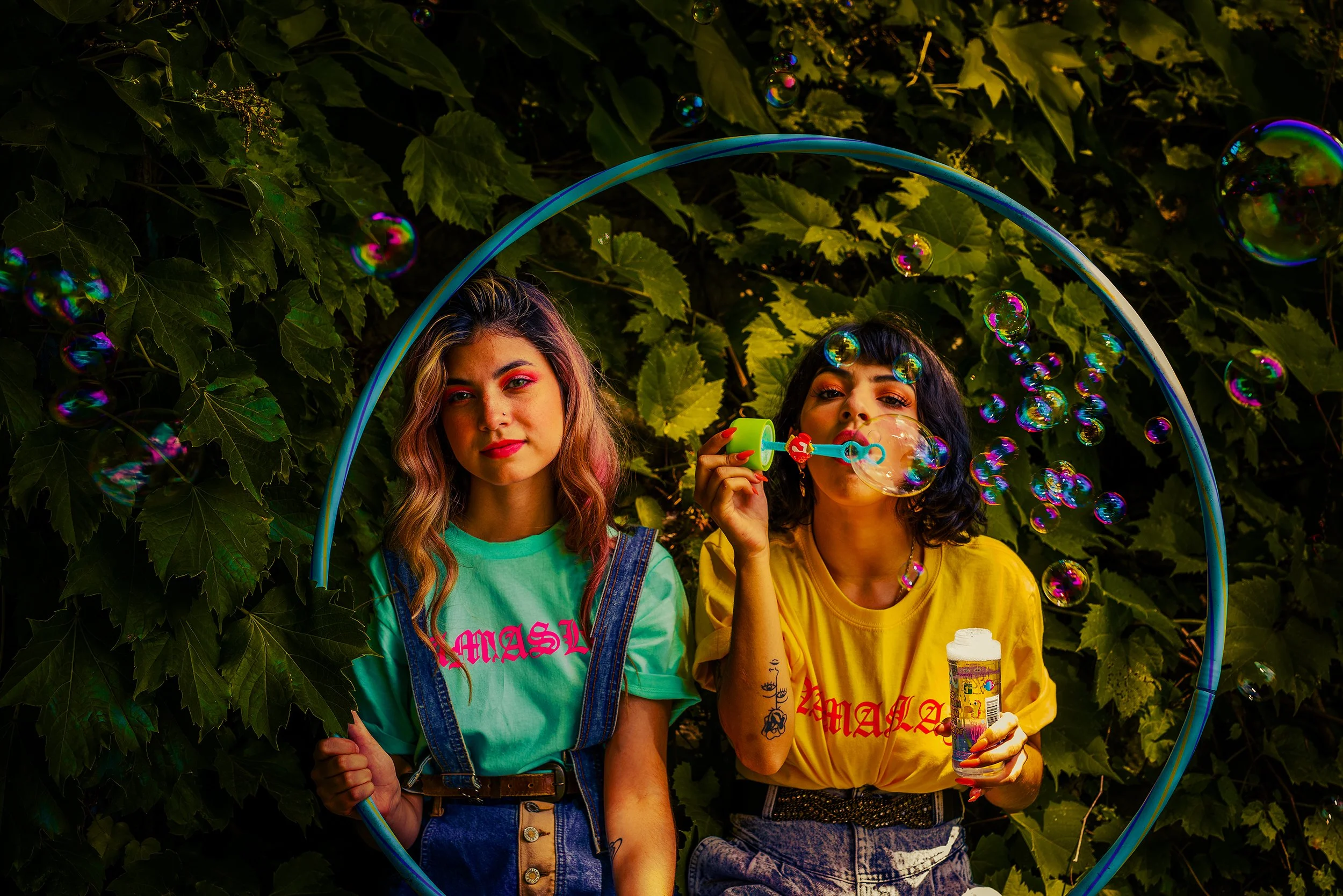 Two young women standing inside a large hula hoop with a backdrop of green leaves. One woman with blonde hair and pink makeup wears a turquoise shirt with pink writing and denim overalls. The other woman with black hair and tattooed arms wears a yell