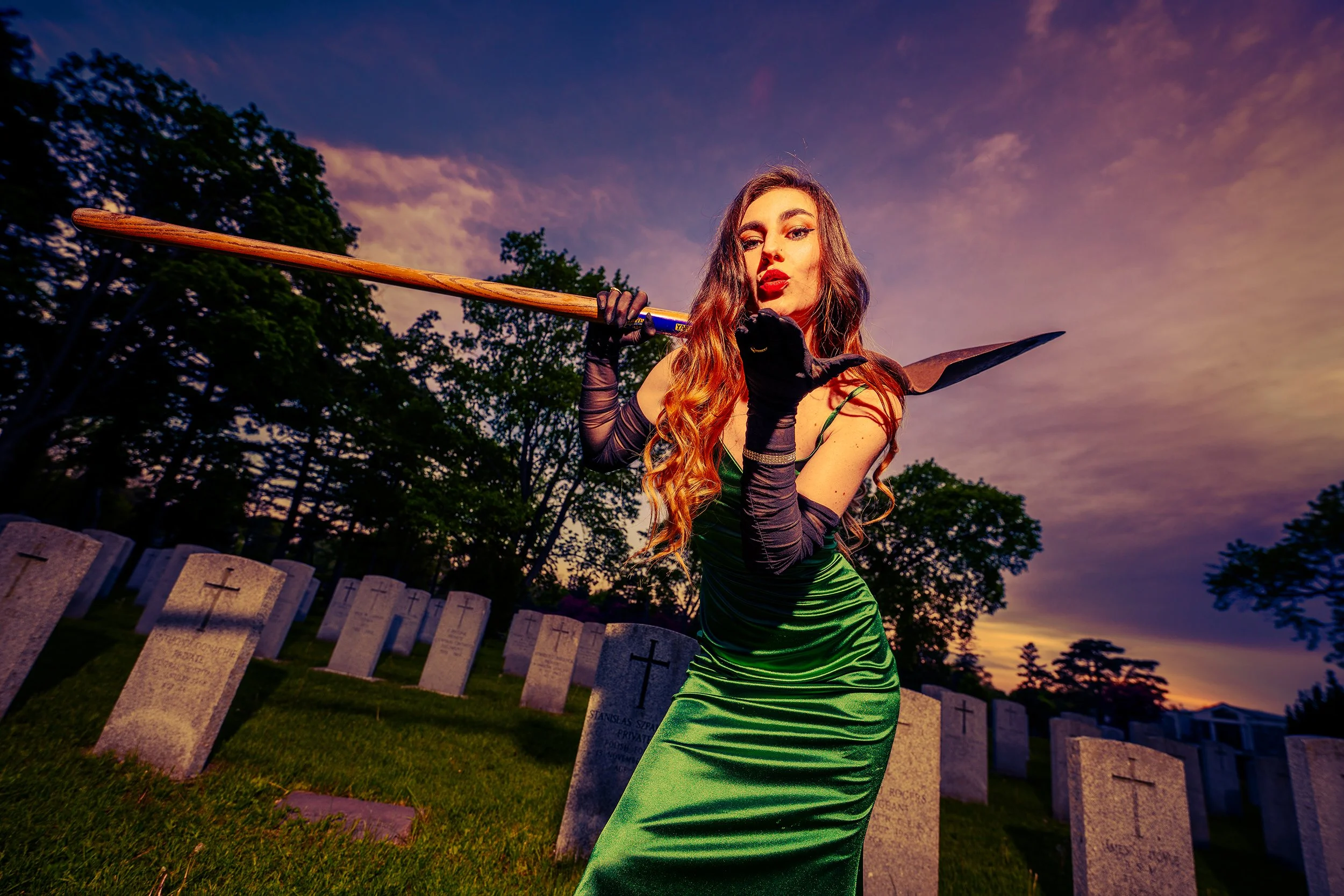 A woman with long of coral-colored hair and bold red lipstick wears a shiny, green, form-fitting dress and black gloves, holding a shovel over her shoulder in a cemetery during sunset.