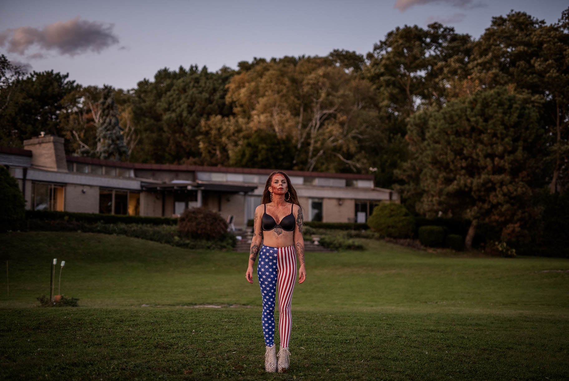A woman with tattoos walking on a grassy lawn during sunset, wearing stars and stripes leggings, a black bra, and beige high-heeled shoes with spiked heels.