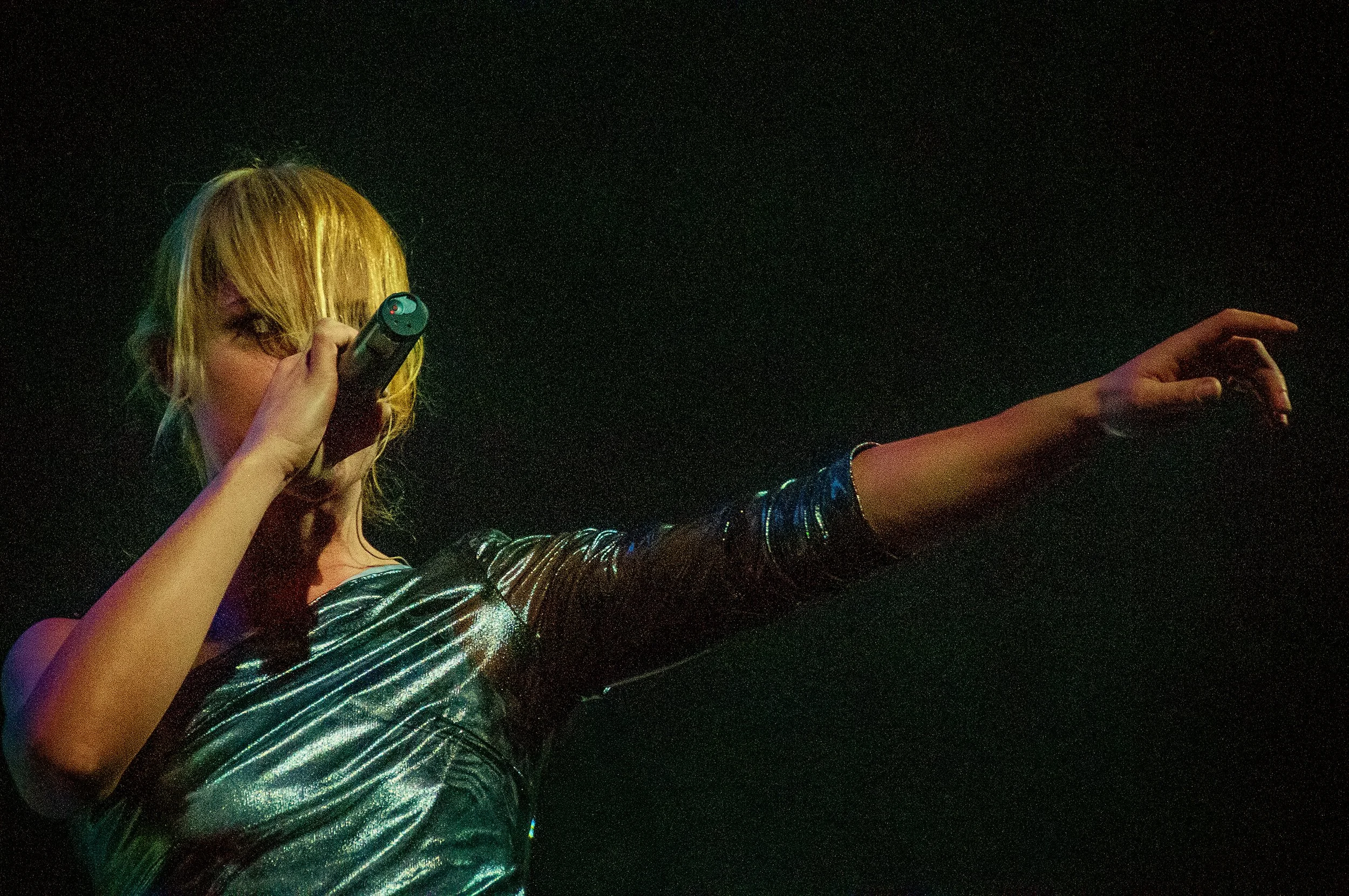 A person with blond hair, wearing a shiny, striped silver outfit, holding a microphone, and extending their other arm against a dark, starry background.