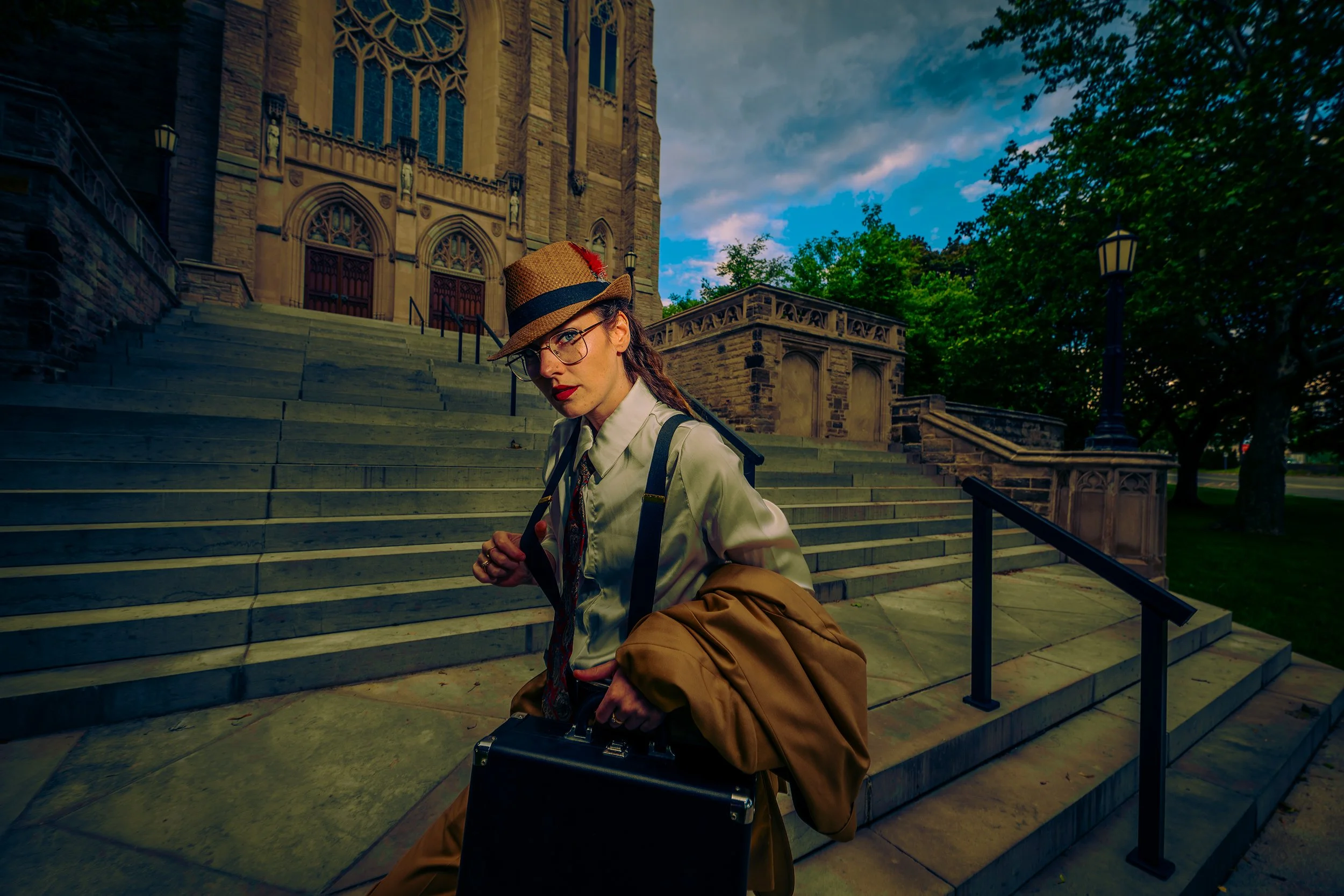 A woman in glasses, a light-colored shirt, and a fedora hat with a red flower, standing on the steps of a historic stone church with gothic architectural features, surrounded by trees on a partly cloudy day.