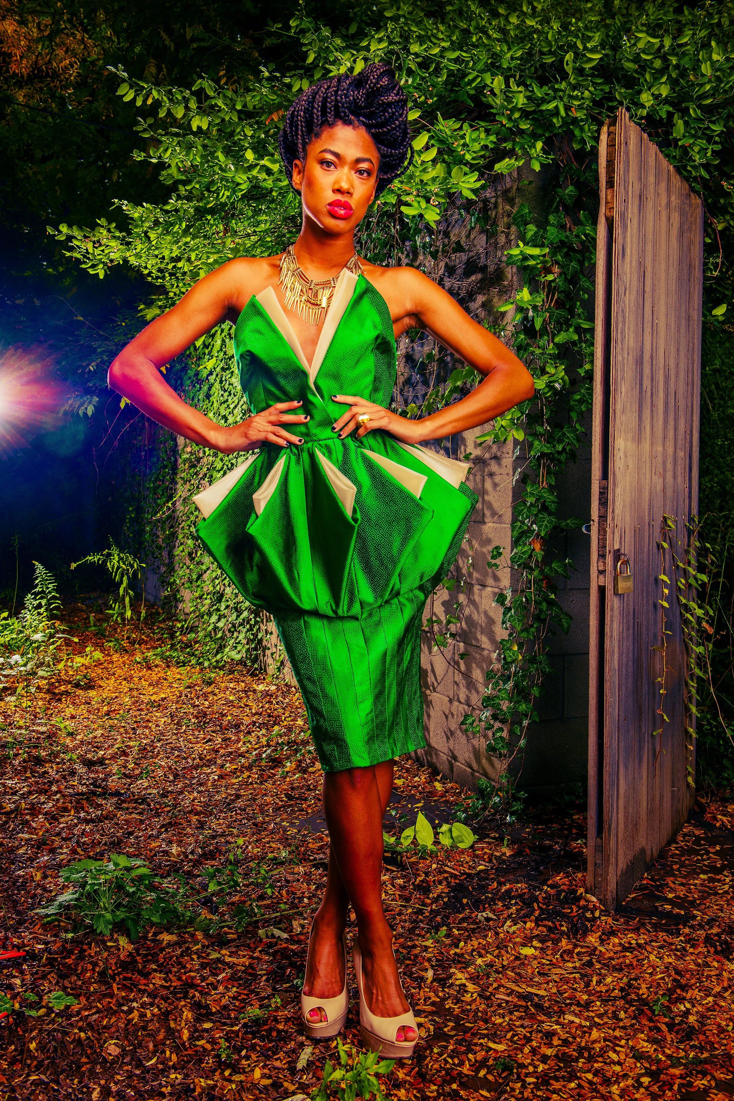 A woman in a vibrant green pleated dress standing outdoors amidst fall foliage, posing with her hands on her hips and wearing beige high heels, gold jewelry, and styled hair.