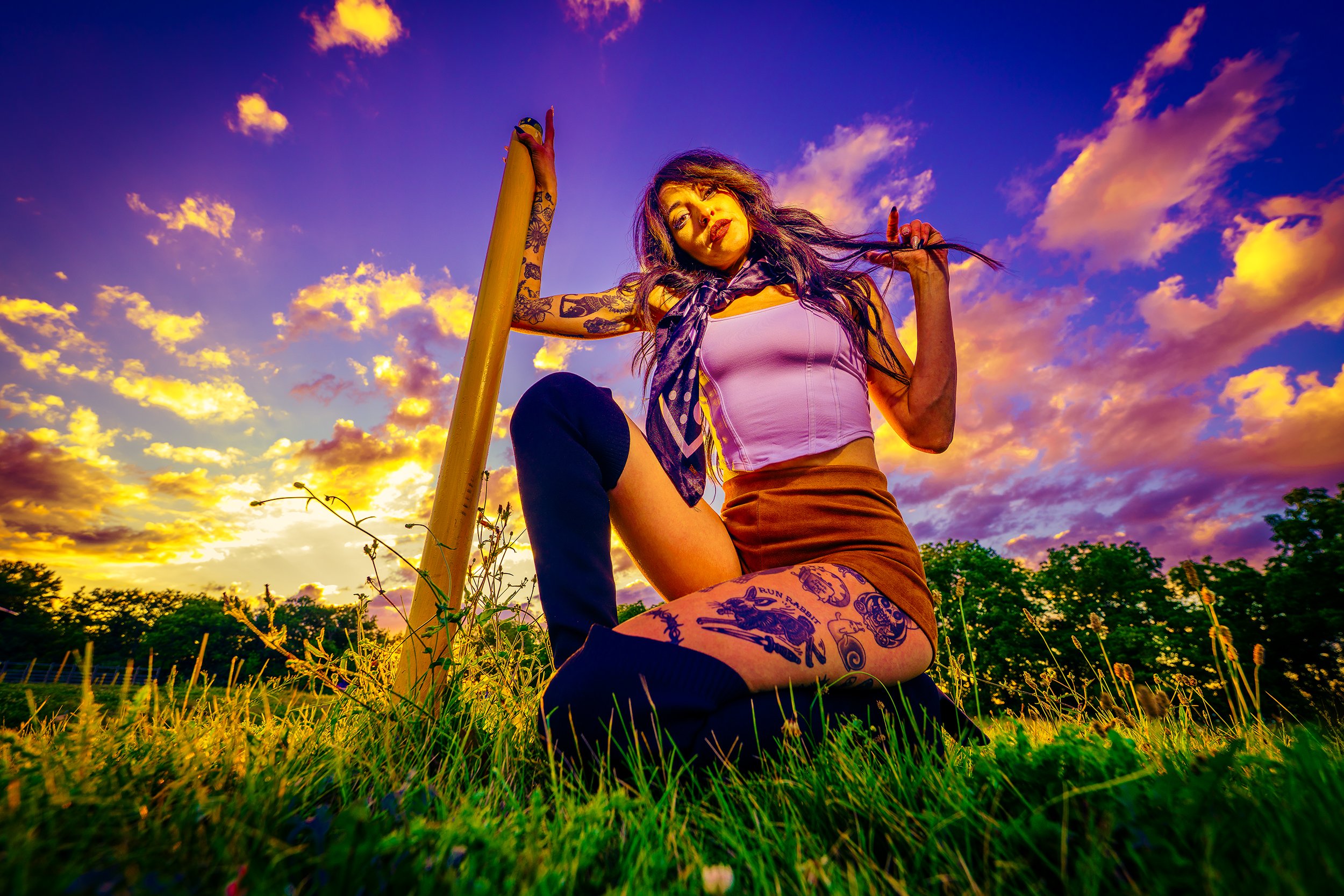 A woman with tattoos, wearing a sleeveless top and skirt, posing outdoors during sunset or sunrise, against a vibrant sky with clouds.