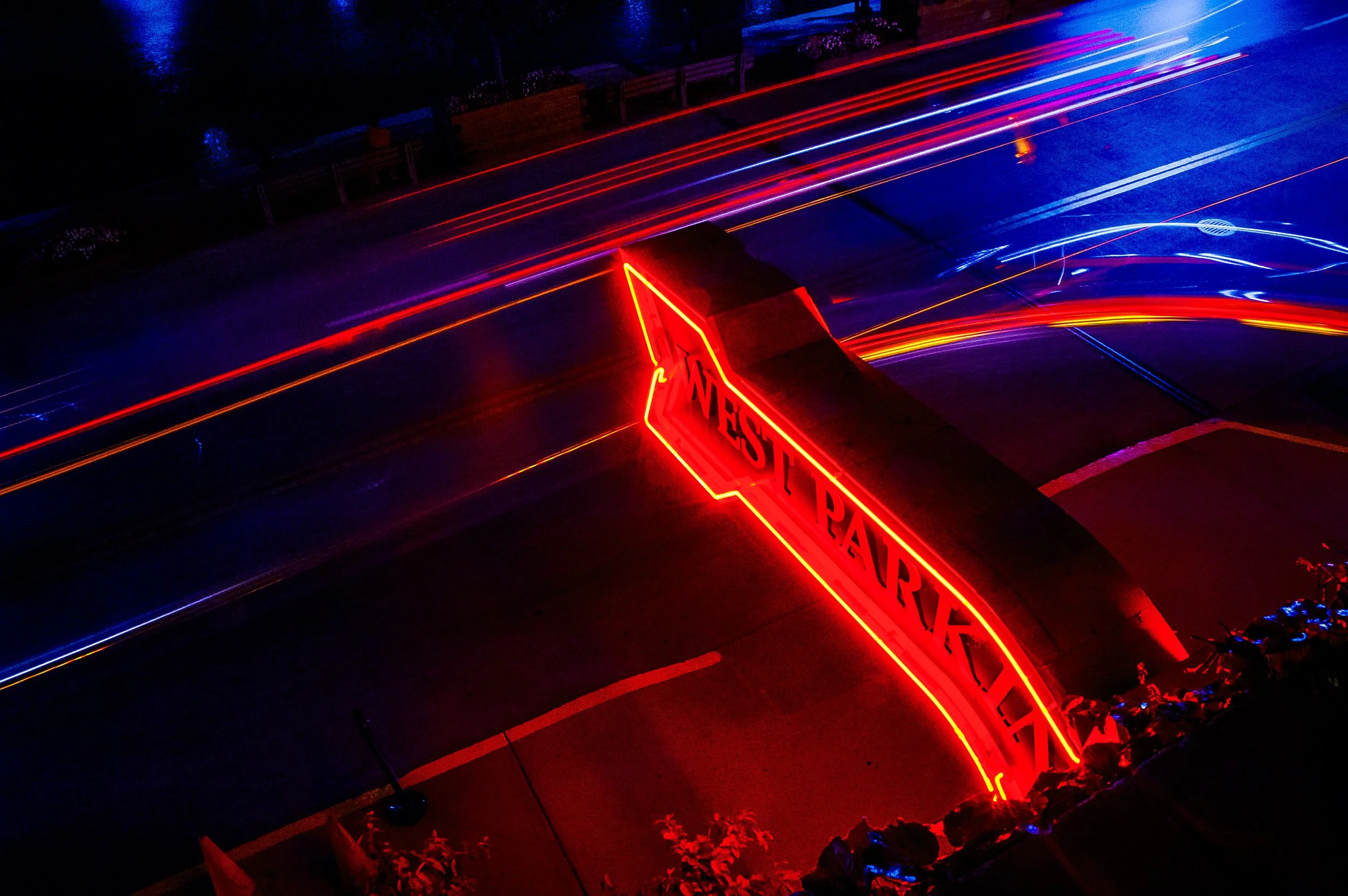 Nighttime view of a decorated parking lot with red and blue lights, a 'Meet Park' illuminated sign, and a row of benches and flower planters along the sidewalk.