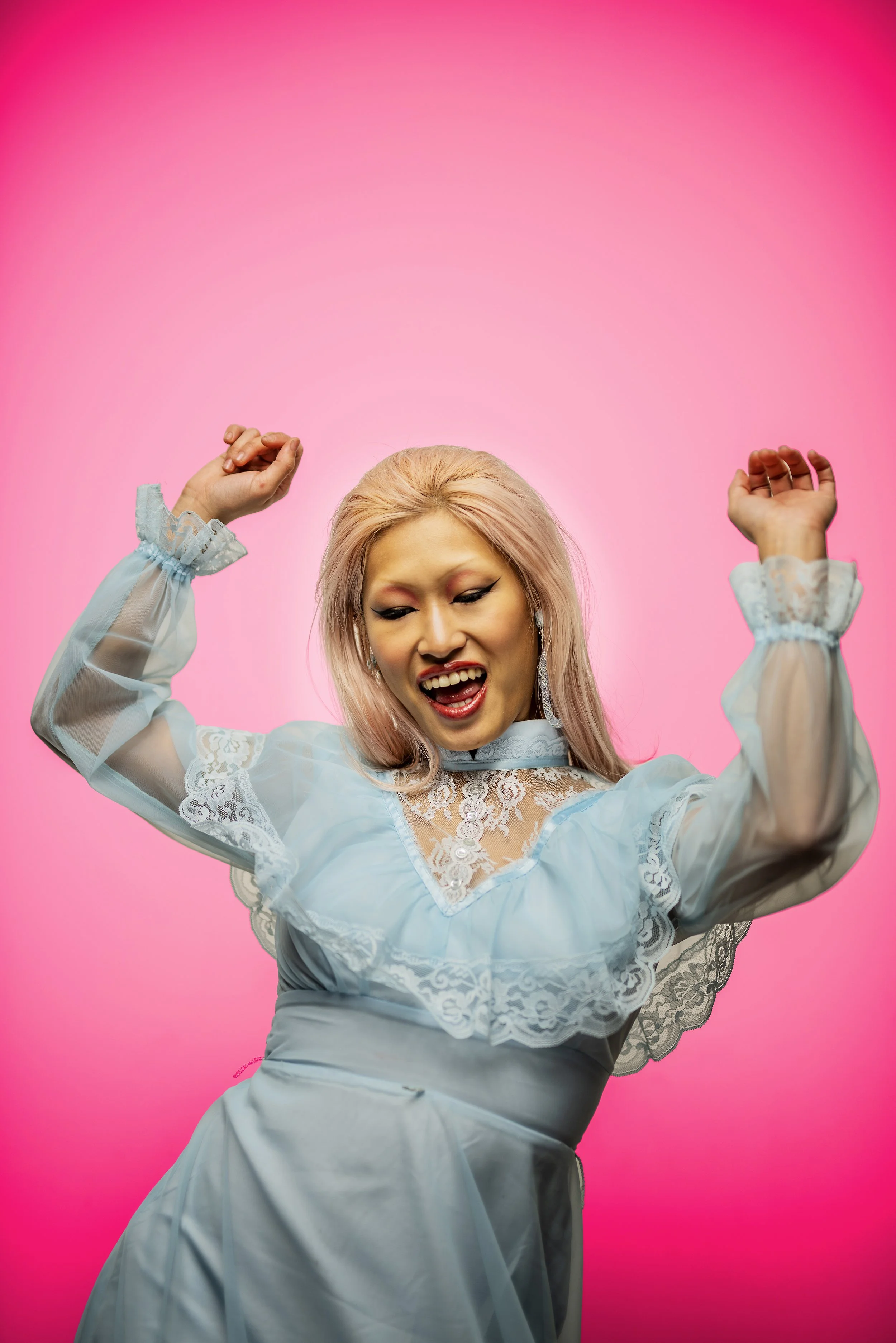 A woman with pink hair, wearing a light blue floral lace dress with sheer sleeves, dancing with her arms raised and smiling against a solid pink background.
