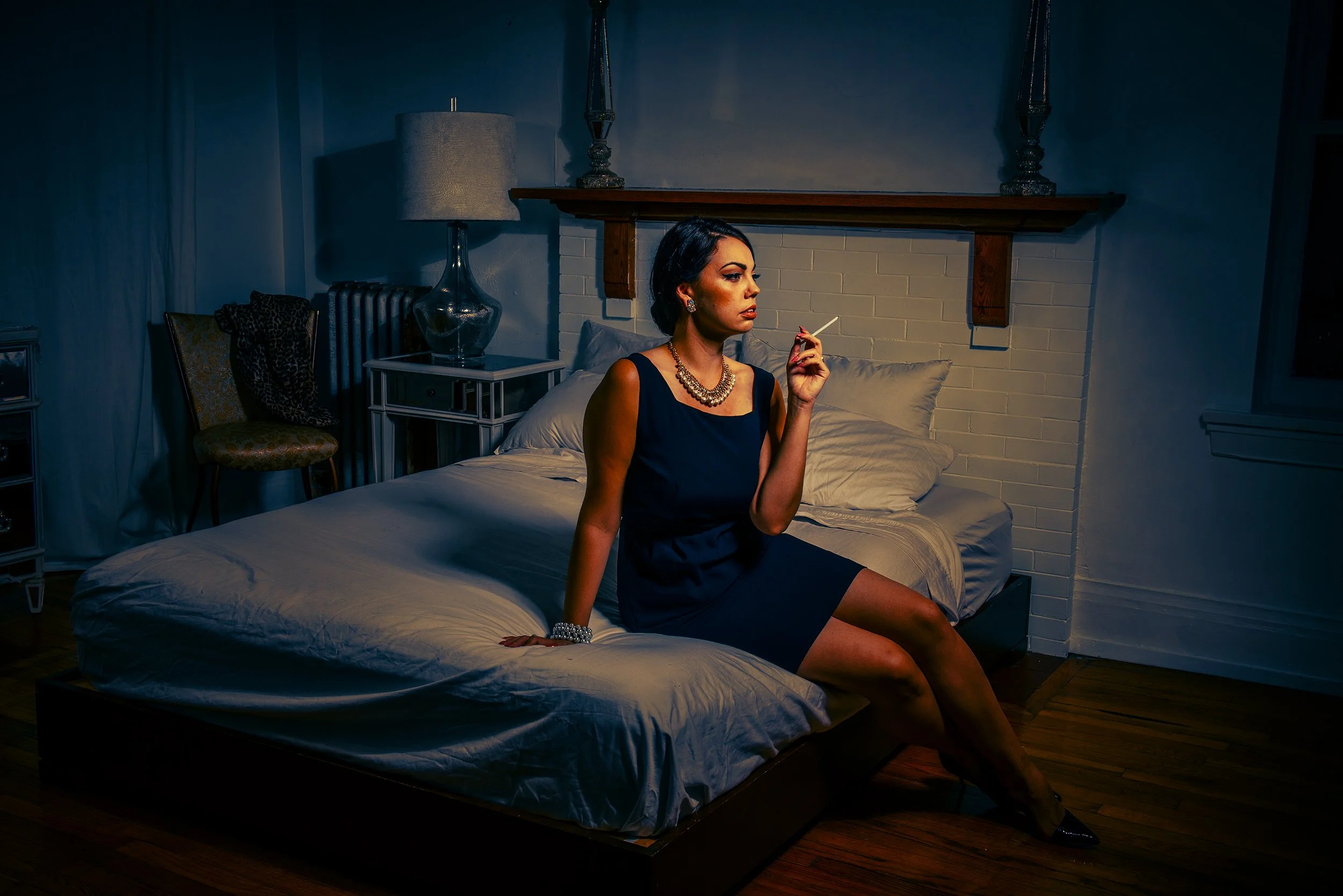 A woman with dark hair, wearing a navy blue dress and jewelry, sitting on the edge of a bed in a dimly lit room, holding a cigarette.