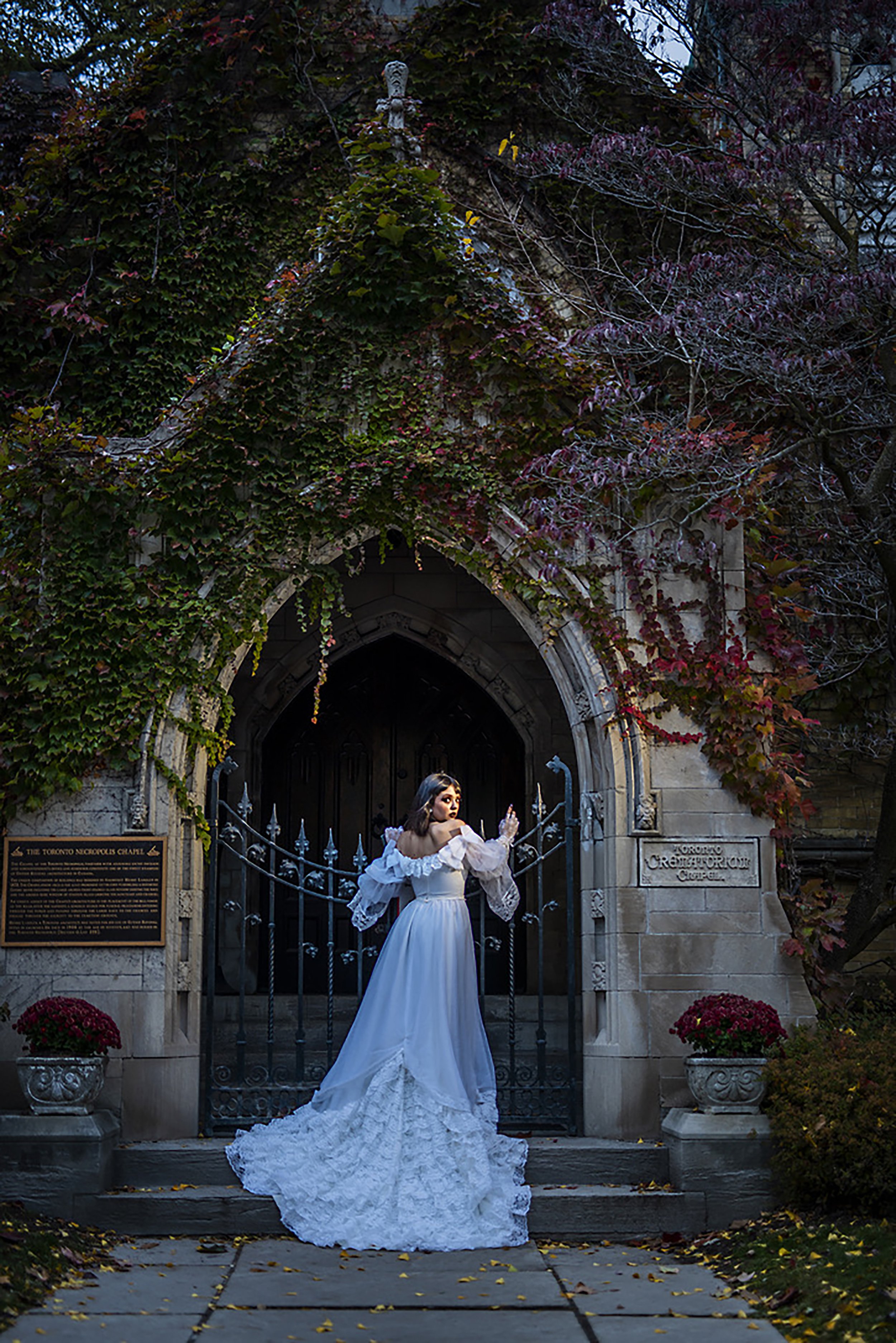 A woman in a white wedding dress with a long train stands in front of an arched, wrought-iron gate covered in ivy and red leaves. She is holding the gate as she looks over her shoulder, with dark hair and a dramatic makeup look. Two stone planters wi