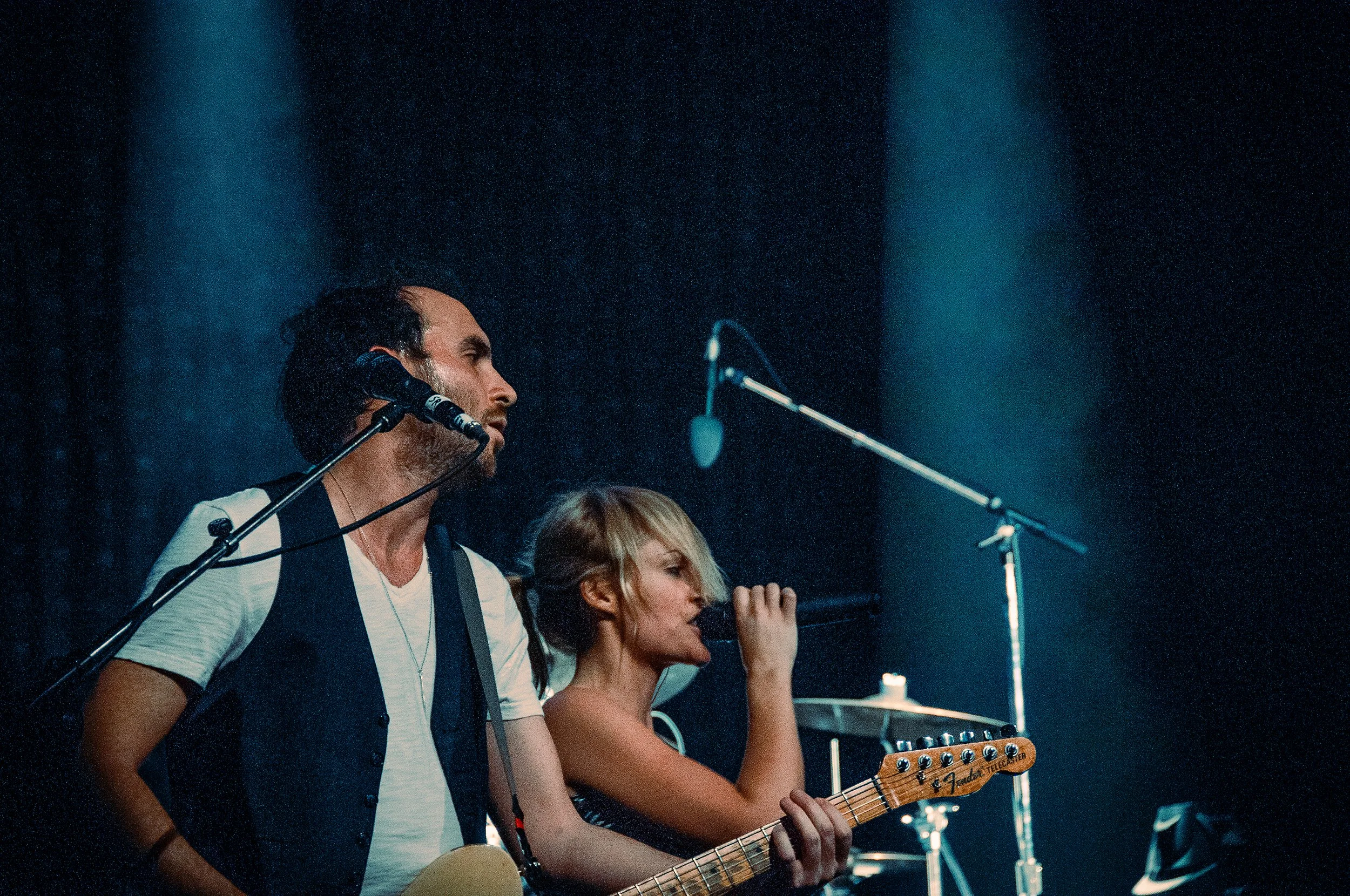 A man playing a guitar and singing into a microphone on stage, with a woman next to him also singing, in a dimly lit concert setting.