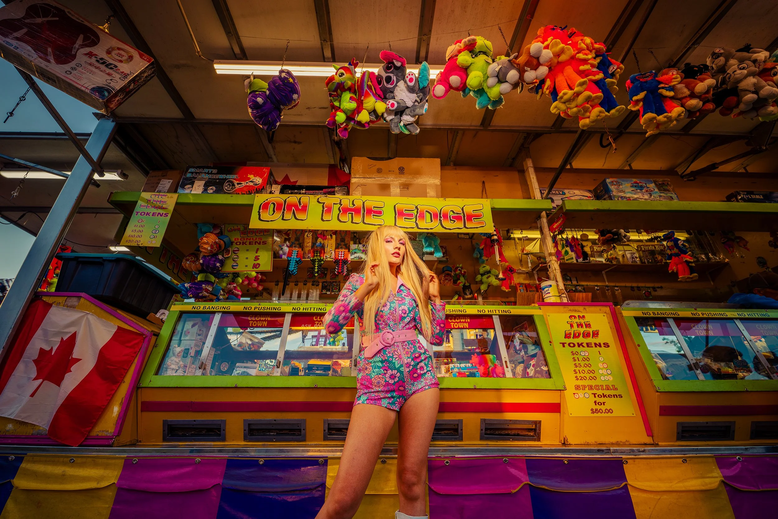 A woman with long blonde hair stands in front of a carnival game booth called 'On The Edge', surrounded by stuffed animal prizes hanging above. The booth features bright colors, a Canadian flag, and various toy prizes and ticket prices displayed on s