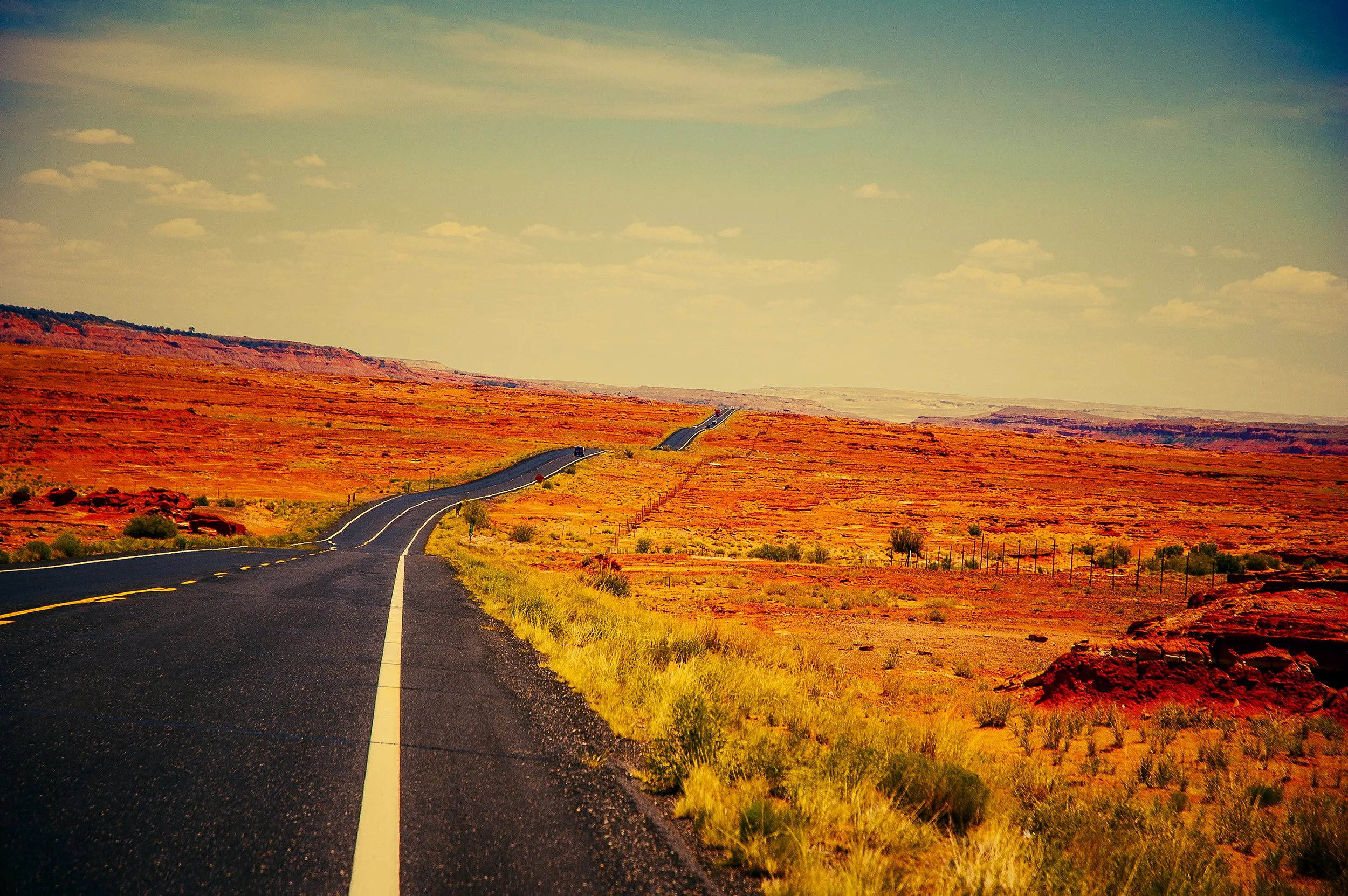 Long winding road through a desert landscape with reddish-orange rocks and sparse vegetation under a partly cloudy sky.