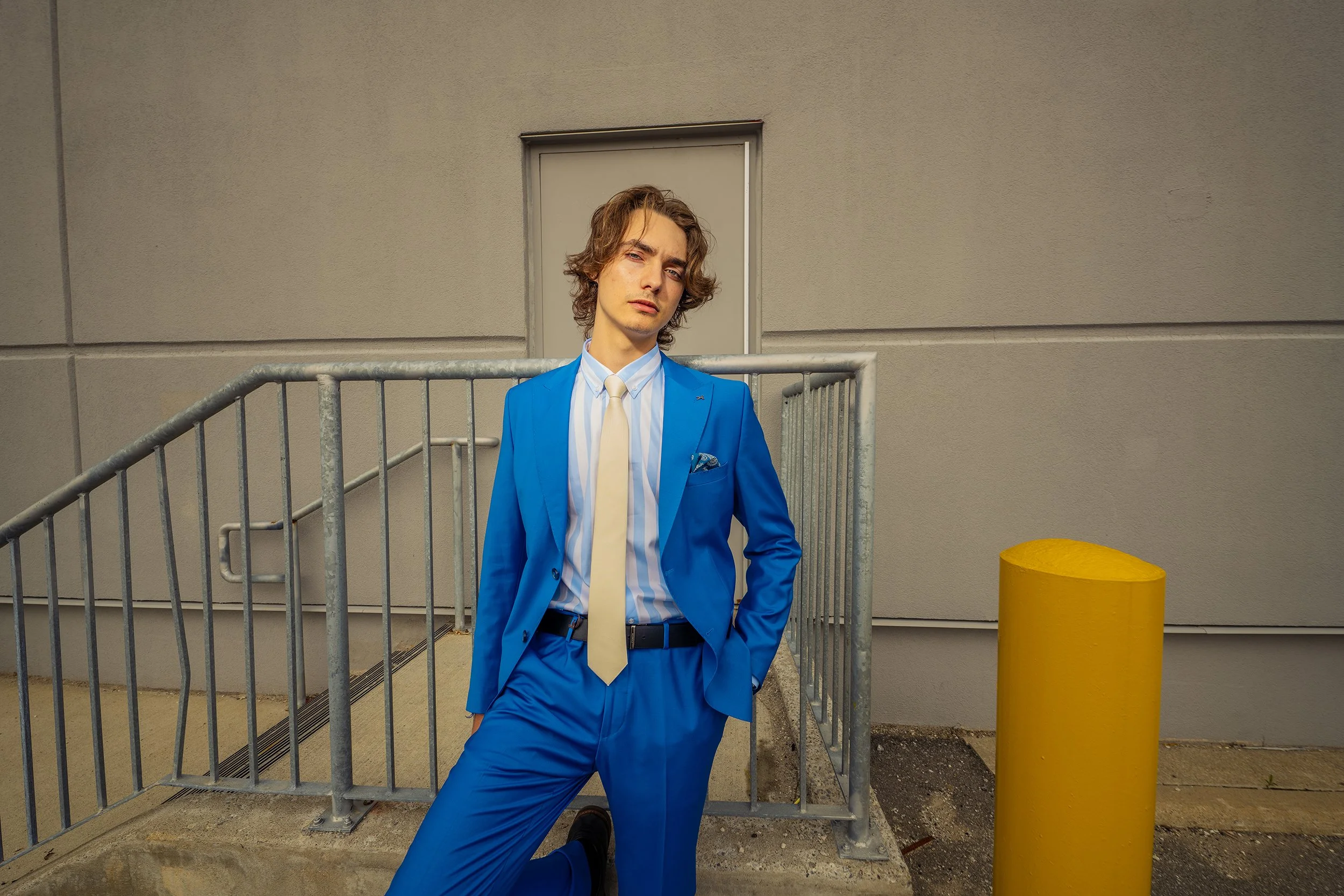 Young man in a blue suit with a white shirt and beige tie standing outdoors near a gray metal railing, with a beige wall and a yellow post in the background.