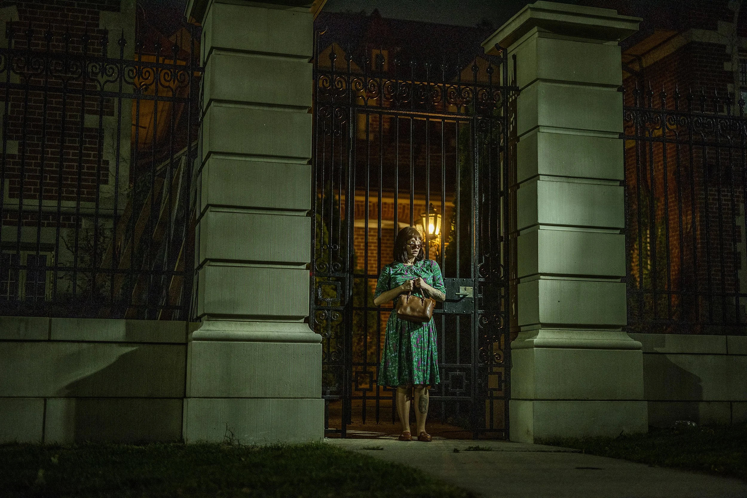 A woman standing in front of a gated entrance at night, illuminated by a nearby light, holding a handbag and wearing glasses and a long, colorful dress.