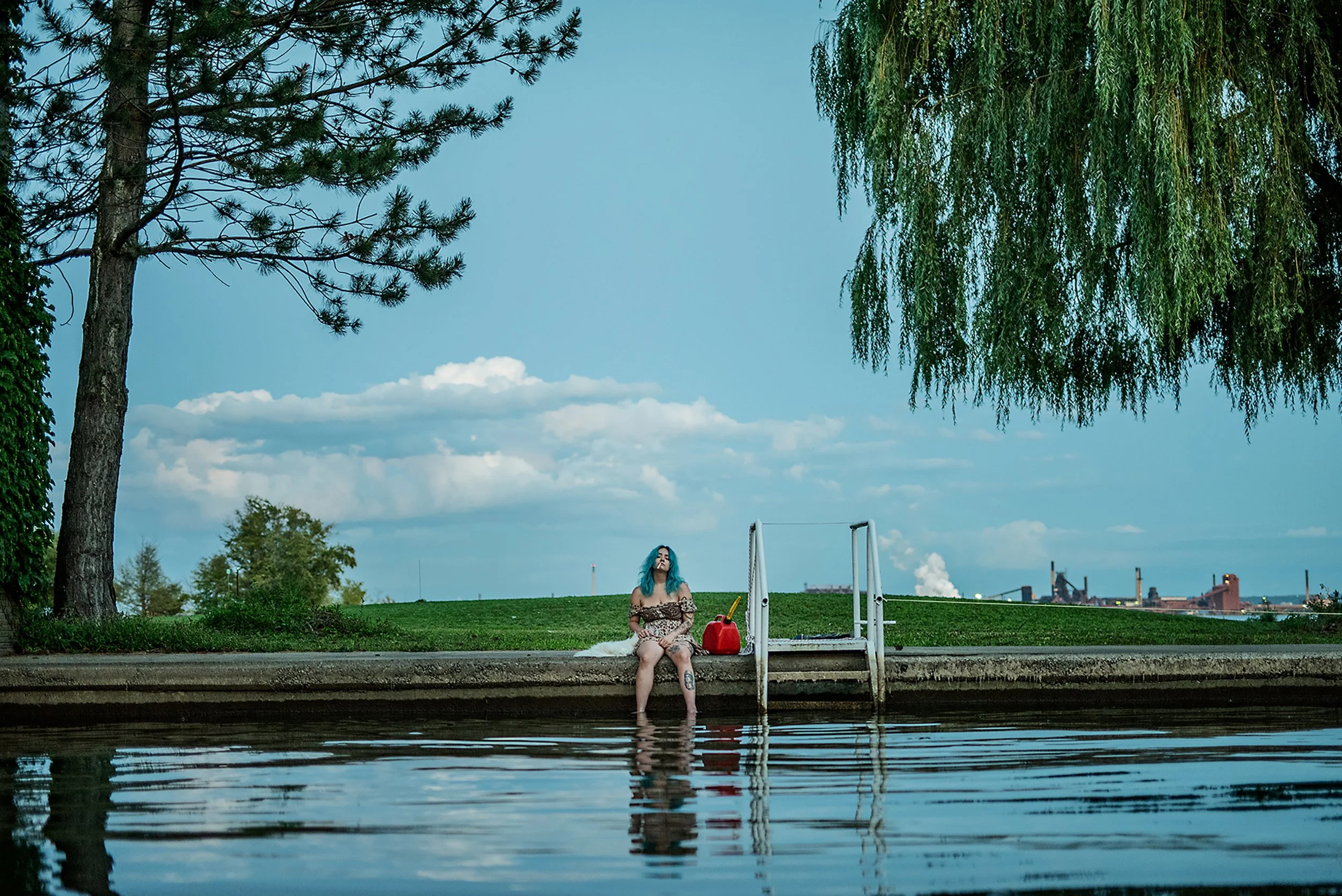 A woman with blue hair sitting by a riverbank on a swimming dock, with a red bag and a yellow toy, surrounded by trees and a cityscape in the background.