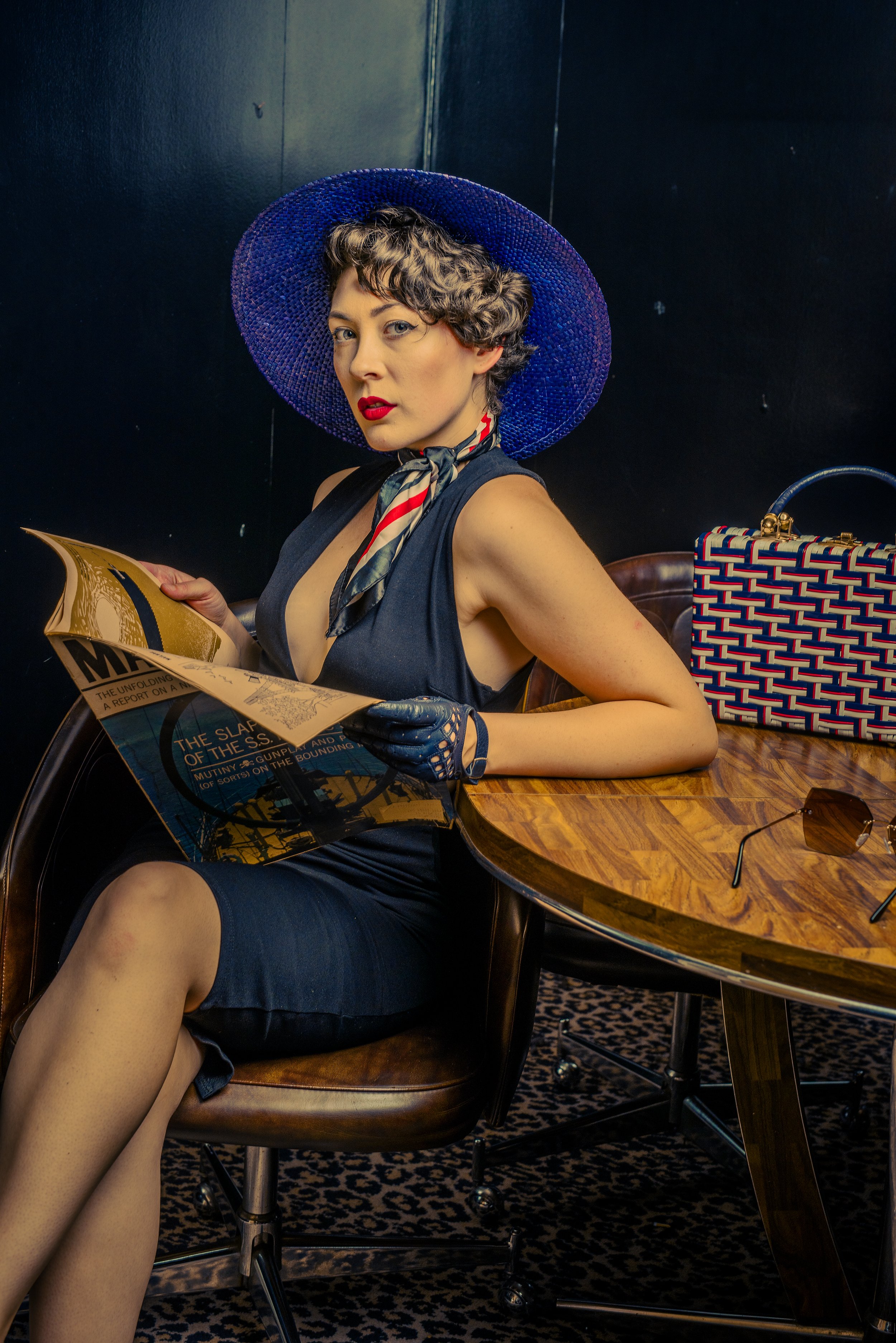 A woman with short curly hair wearing a large blue sun hat, red lipstick, and a navy sleeveless dress, sitting at a wooden table with a patterned handbag and sunglasses, reading a newspaper against a dark wall background.