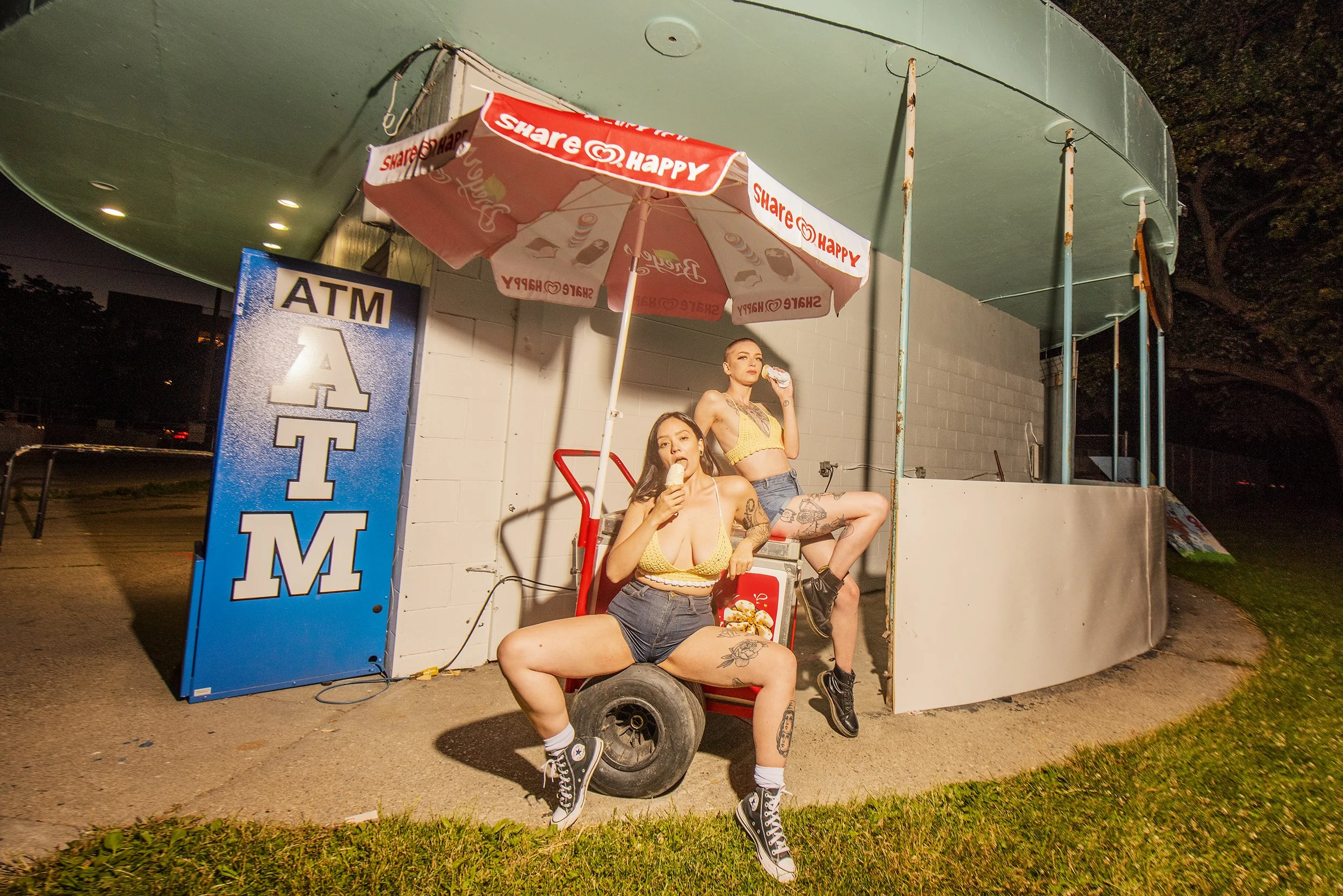 Two women with tattoos in yellow tops and denim shorts, eating ice cream outside a small, rounded, green and gray building at night. One woman is sitting on a small red cart, the other is sitting on the edge of the building's railing. A large blue an