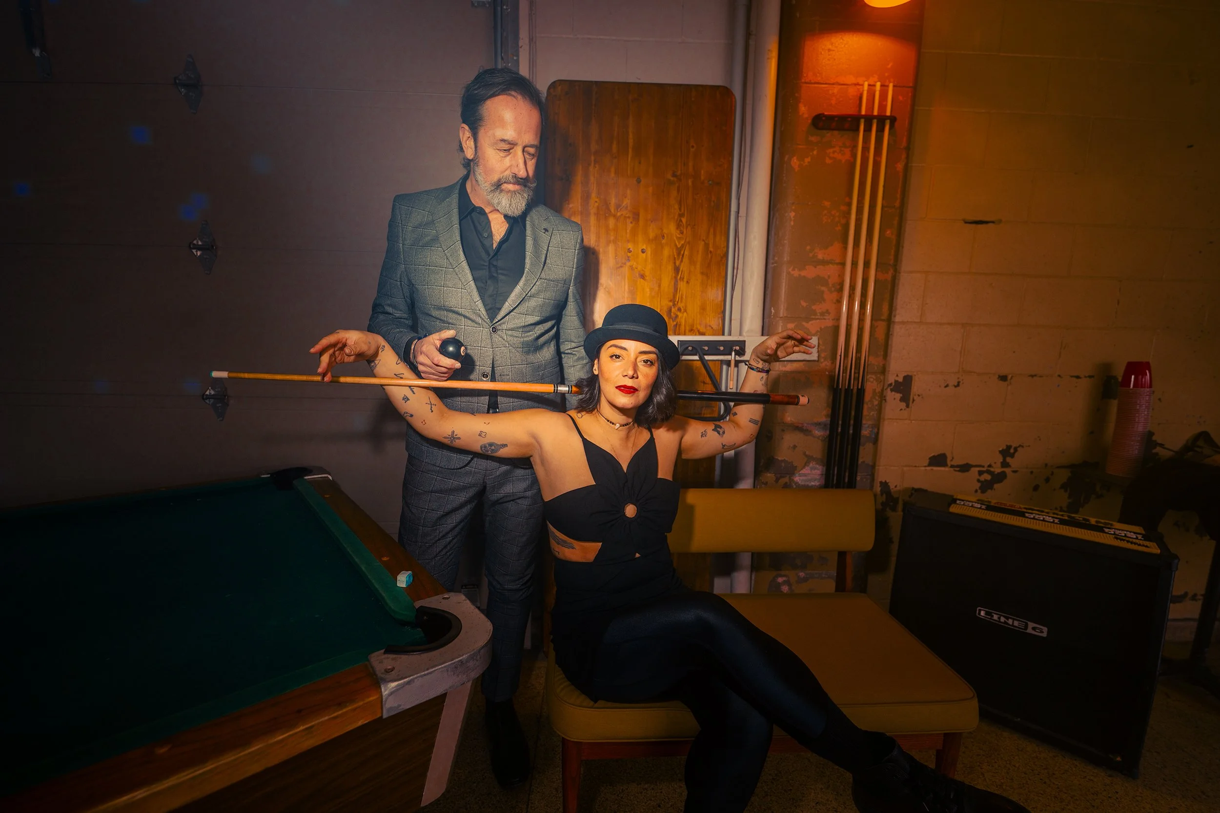 A woman dressed in black with tattoos and a black hat sits on a yellow chair, holding a pool cue horizontally with her arms raised. A man in a checkered blazer stands behind her, holding a small object, and appears to be overseeing her pose in a diml