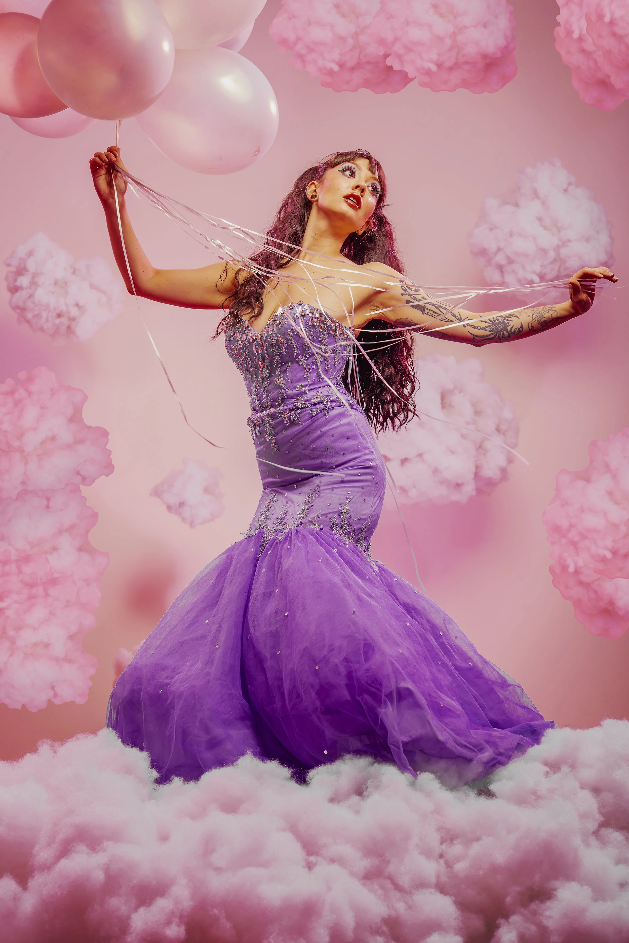 A woman in a purple gown holding pink balloons in a cloud-filled, pink, dreamy setting.