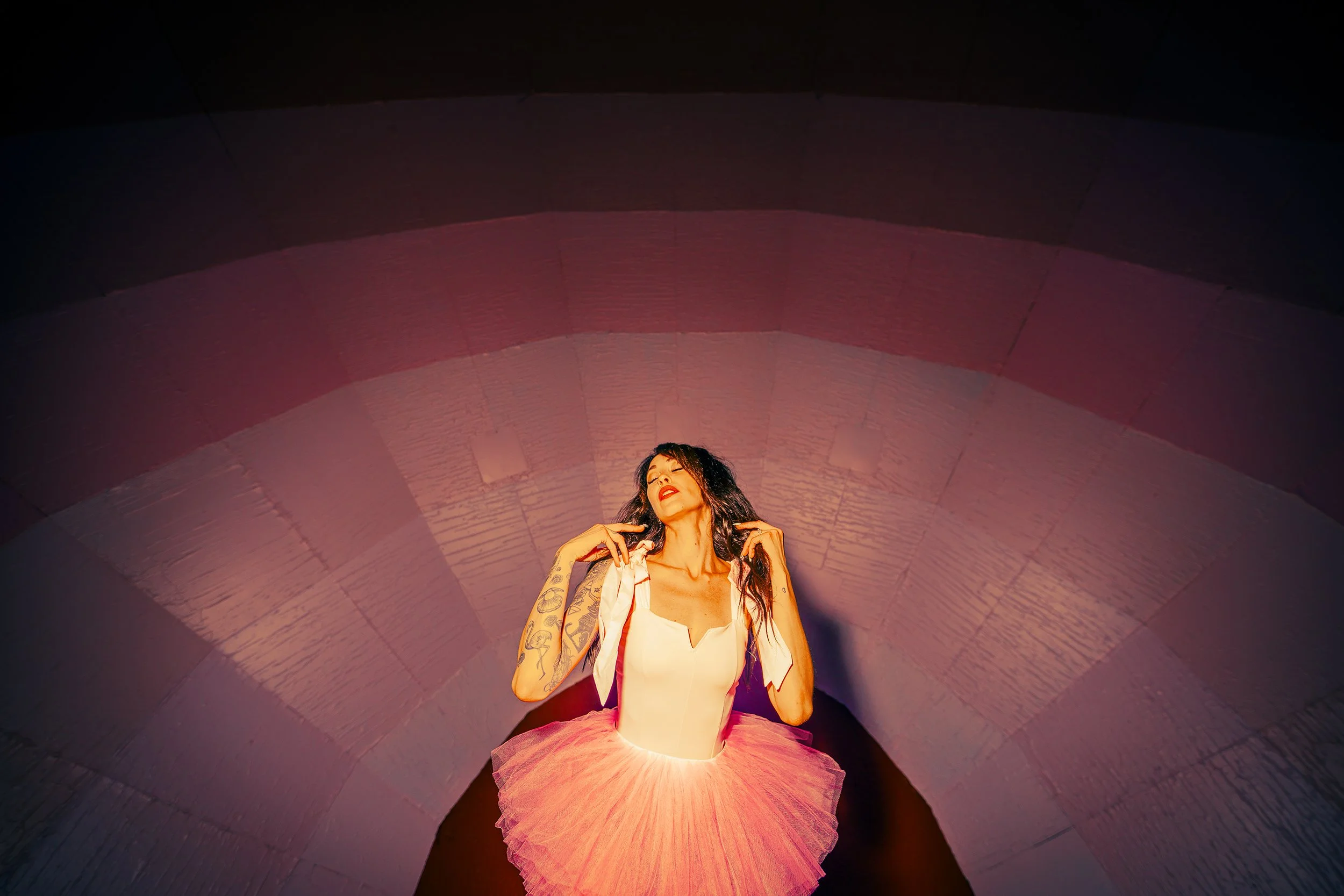 A woman with dark hair and tattoos on her arms, wearing a white top and pink tutu, sitting inside a large, semi-circular, metallic tunnel with pink lighting.